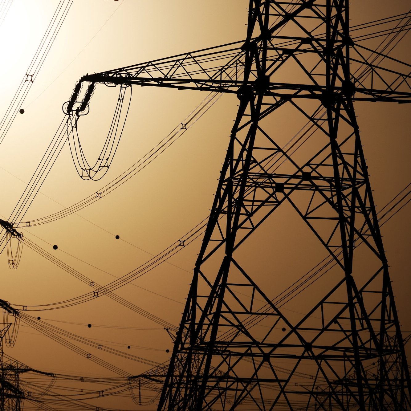 Silhouettes of tall electrical towers against a hazy, orange sunset sky, with power lines connecting them.
