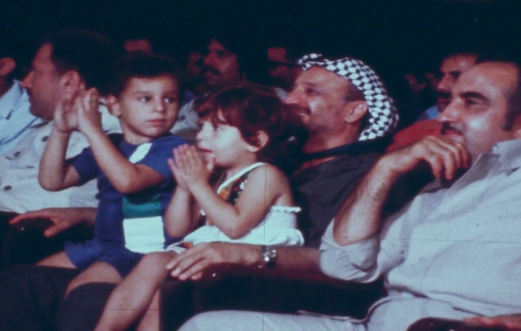 La imagen muestra un grupo de personas sentadas en un evento, aplaudiendo. En el centro de la imagen, hay dos niños: un niño que lleva una camiseta de colores y una niña que lleva un vestido claro. Detrás de ellos, se puede ver a un hombre con una keffiyeh (un pañuelo típicamente árabe) y otros asistentes en el fondo. La atmósfera parece festiva, y todos están disfrutando del momento.