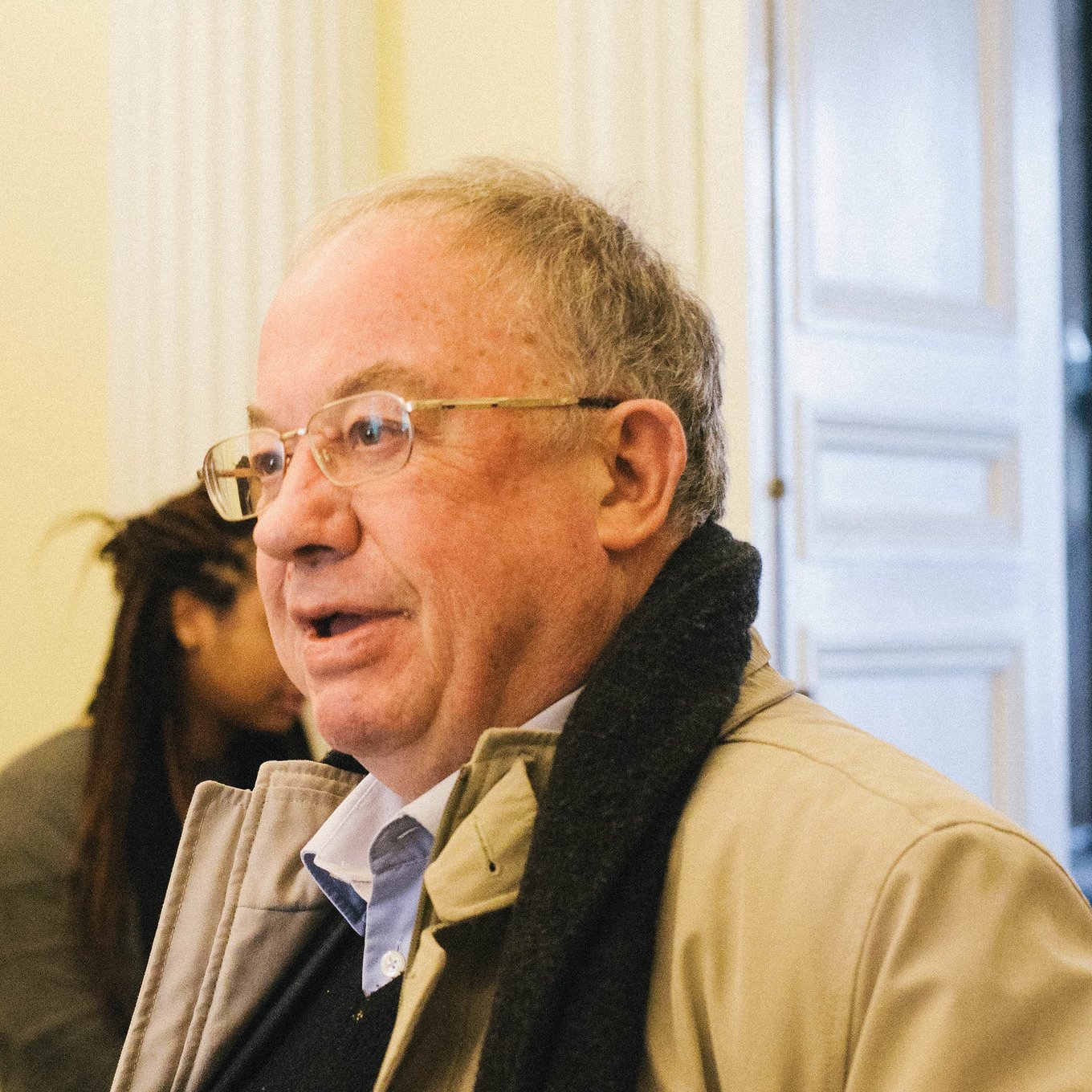 A man with glasses, light hair, and a tan coat stands indoors, speaking with someone behind him.