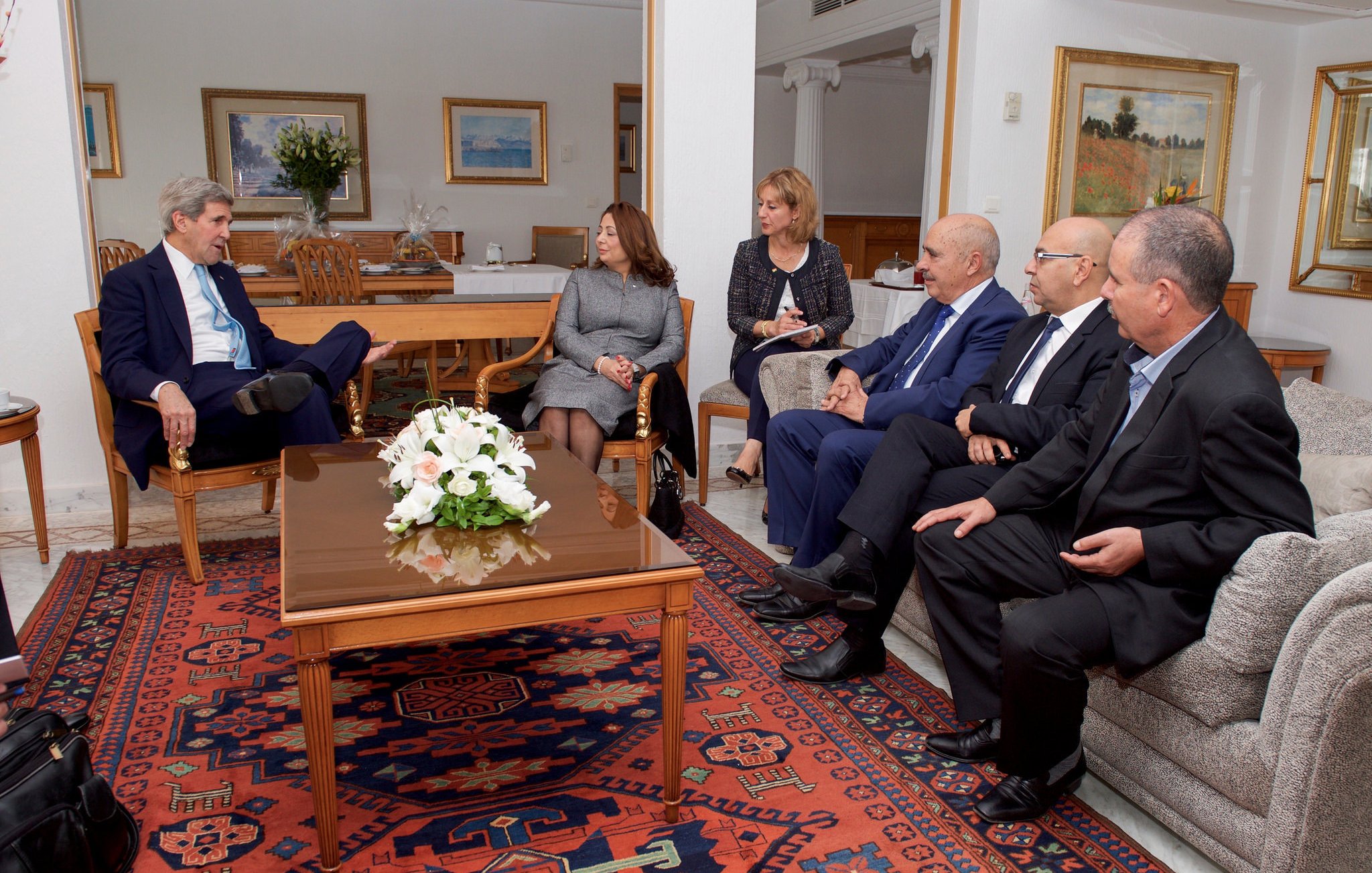 تظهر الصورة مجموعة من الأشخاص يجلسون في غرفة معيشة أنيقة. في المنتصف، يوجد طاولة قهوة تحمل زهورًا بيضاء. يبدو أن هناك نقاشًا أو اجتماعًا يجري بينهم. يجلس شخص يرتدي بدلة زرقاء في الجهة اليسرى، بينما يجلس الآخرون على الأرائك المحيطة. الخلفية تحتوي على لوحات فنية ونباتات، مما يضفي جوًا مريحًا على المكان.