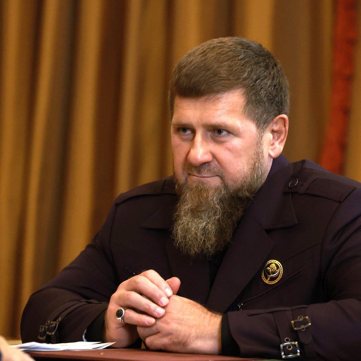Homme avec une barbe, vêtu d'un costume sombre, assis à une table.