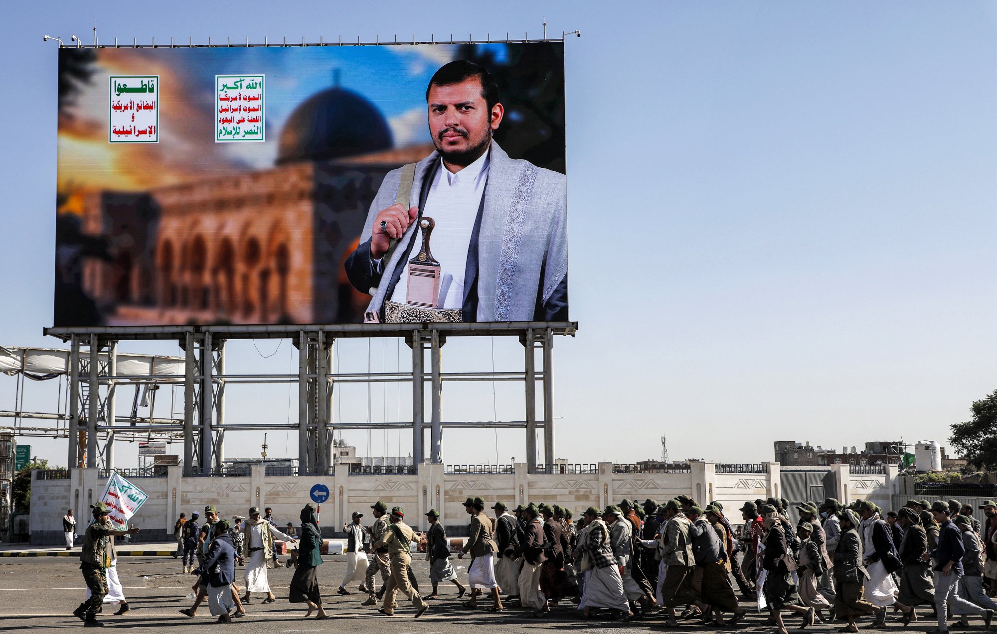L'image montre un grand panneau d'affichage avec un portrait d'un homme en tenue traditionnelle, probablement un leader ou une figure politique, devant un paysage architectural. En dessous, un groupe de personnes marche en procession, portant des drapeaux et des affiches. L'environnement semble urbain, suggérant une manifestation ou un événement public. Les écriteaux sur le panneau en arabe ajoutent au contexte culturel et politique de l'image.