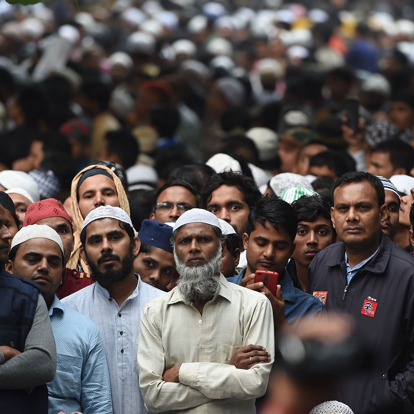 L'image montre une grande foule d'hommes, principalement vêtus de vêtements traditionnels, avec plusieurs d'entre eux portant des couvre-chefs. Les expressions des gens varient, certains semblent attentifs, tandis que d'autres regardent vers l'avant. L'ambiance dégage un sentiment de concentration et de rassemblement, possiblement pour un événement ou une cérémonie.