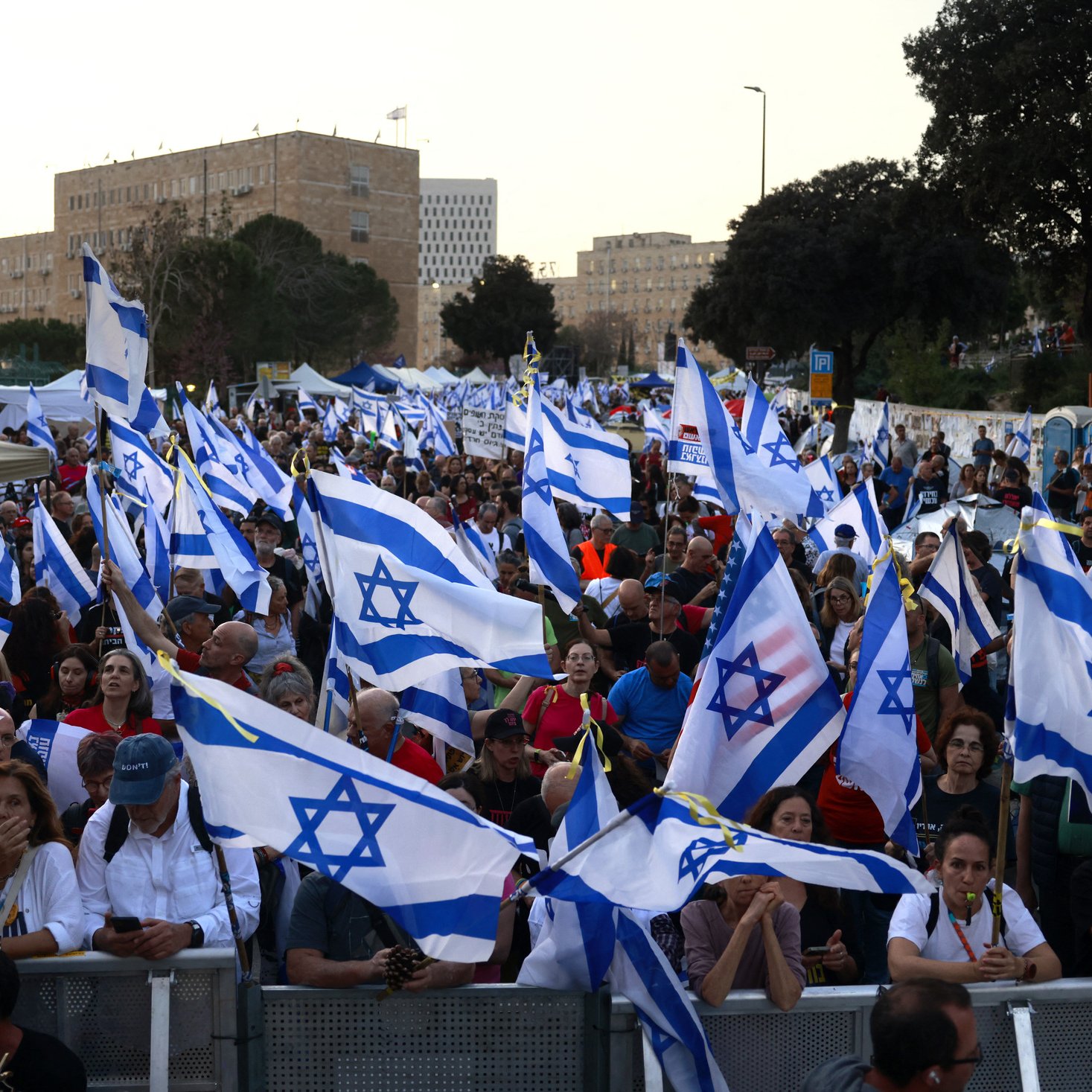 L'image montre une grande foule de personnes rassemblées, brandissant des drapeaux israéliens. Les manifestants semblent réunis pour une occasion particulière, avec une ambiance dynamique. En arrière-plan, on peut apercevoir des bâtiments et des structures urbaines, tandis que des tentes sont installées, peut-être pour fournir des services aux participants. L'ensemble illustre une manifestation significative, avec une forte présence de la couleur bleue et blanche des drapeaux.