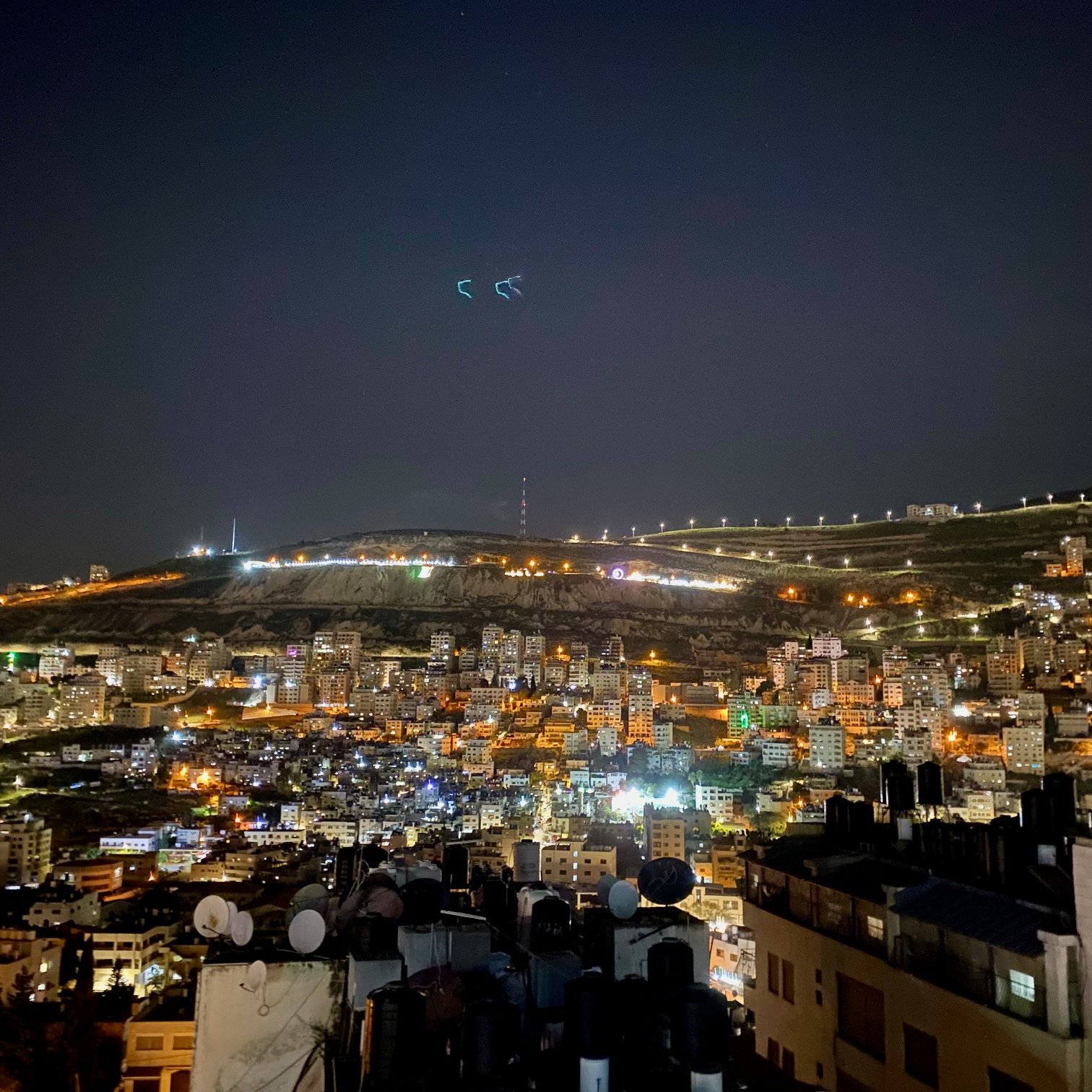 L'image montre une vue nocturne d'une ville illuminée. On peut voir de nombreux bâtiments agencés en terrasses, avec des lumières qui scintillent dans l'obscurité. À l'arrière-plan, des collines sont éclairées, et quelques structures sont visibles au sommet. L'atmosphère est tranquille, avec un ciel nocturne clair. Les antennes et structures en premier plan ajoutent une dimension urbaine à cette scène paisible.