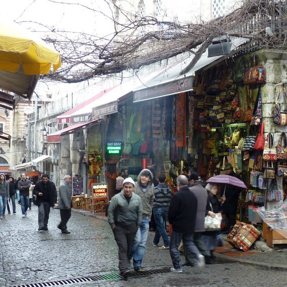 L'image montre une ruelle animée d'un marché. Les pavés sont humides, suggérant peut-être qu'il a récemment plu. Les étals sont remplis de sacs colorés et d'autres articles artisanaux, donnant une impression de vivacité et de diversité. Des gens se déplacent dans la rue, certains se protégeant sous des parapluies. Au-dessus, des branches nues d'arbres créent une atmosphère presque nostalgique, tandis que des auvents jaunes ajoutent une touche de couleur à la scène.