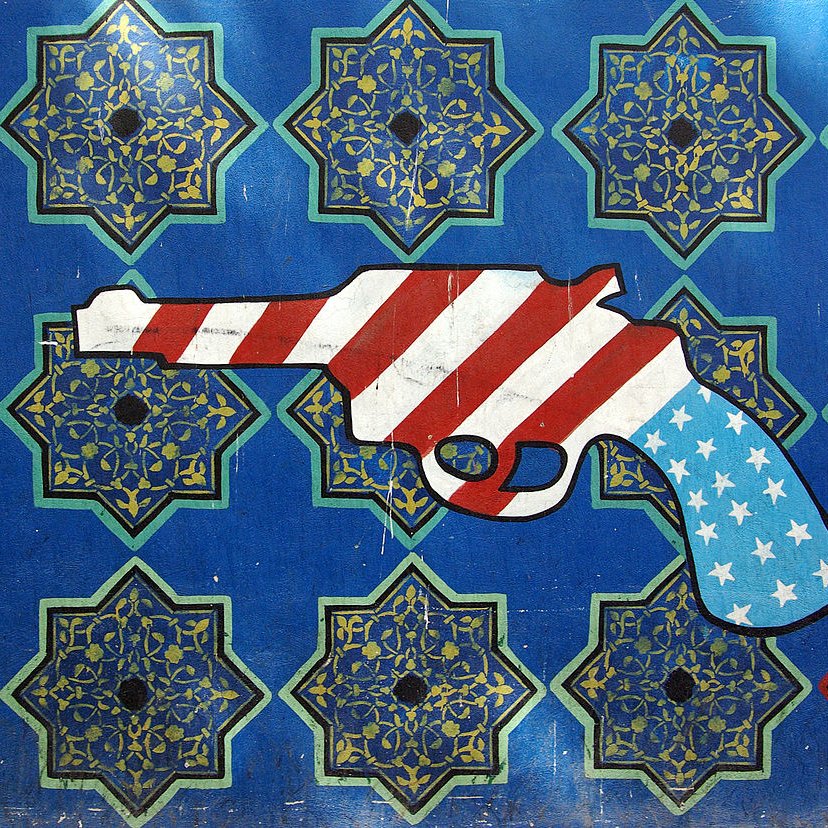 L'image représente un pistolet stylisé recouvert des motifs du drapeau américain, avec des rayures rouges et blanches et des étoiles blanches sur un fond bleu. Le pistolet est placé sur un arrière-plan orné de motifs géométriques complexes aux couleurs vives, qui évoquent des designs traditionnels. Le contraste entre le pistolet et l'arrière-plan artistique souligne un message potentiellement provocateur.