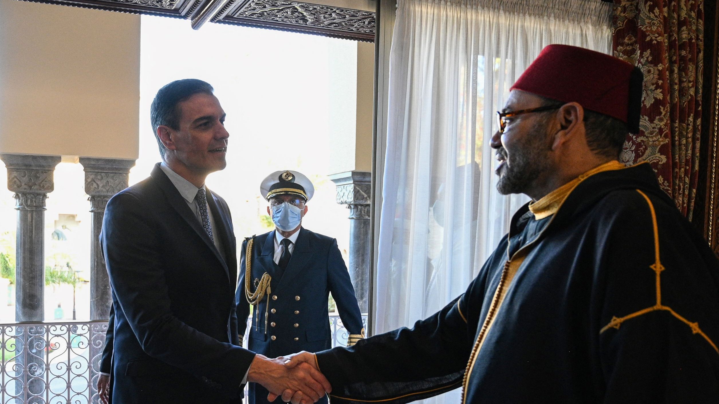 تظهر في الصورة لقاء بين شخصين، حيث يقوم أحدهما بمصافحة الآخر. يبدو أن الاجتماع رسمي، وذلك بسبب الملابس الأنيقة التي يرتديها الشخصان. في الخلفية، يوجد شخص ثالث يبدو أنه يؤدي دورًا رسميًا أيضًا. الأجواء توحي بالاحترام والتقدير بين الحضور. الأثاث وإعداد الغرفة يظهران بشكل تقليدي وفاخر.