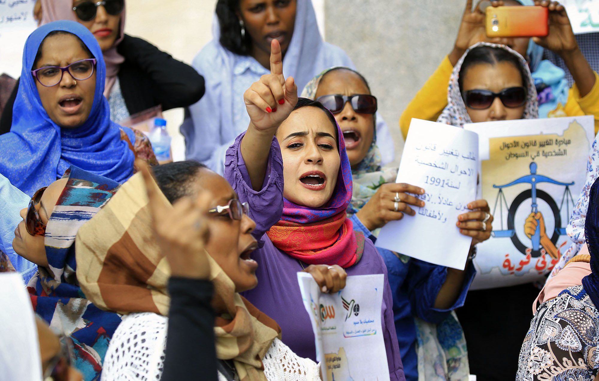 L'image montre un groupe de femmes lors d'une manifestation. Elles portent des foulards et des vêtements colorés. Certaines personnes lèvent les mains et expriment des slogans avec une grande énergie. Des pancartes sont visibles, portant des messages concernant des droits ou des revendications sociales. L'ambiance est dynamique et engagée, avec un fort esprit de solidarité parmi les participantes.