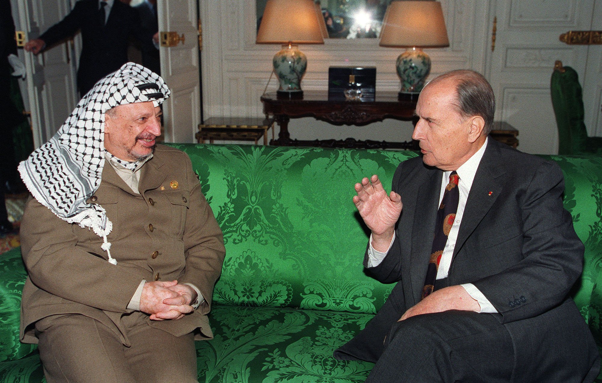 L'image montre deux hommes assis sur un canapé vert décoré. L'un des hommes porte un costume beige avec un keffieh, tandis que l'autre est en costume sombre avec une cravate à motifs. Ils semblent engagés dans une conversation animée. En arrière-plan, on aperçoit des éléments de mobilier et une lumière douce, suggérant un cadre intérieur élégant.