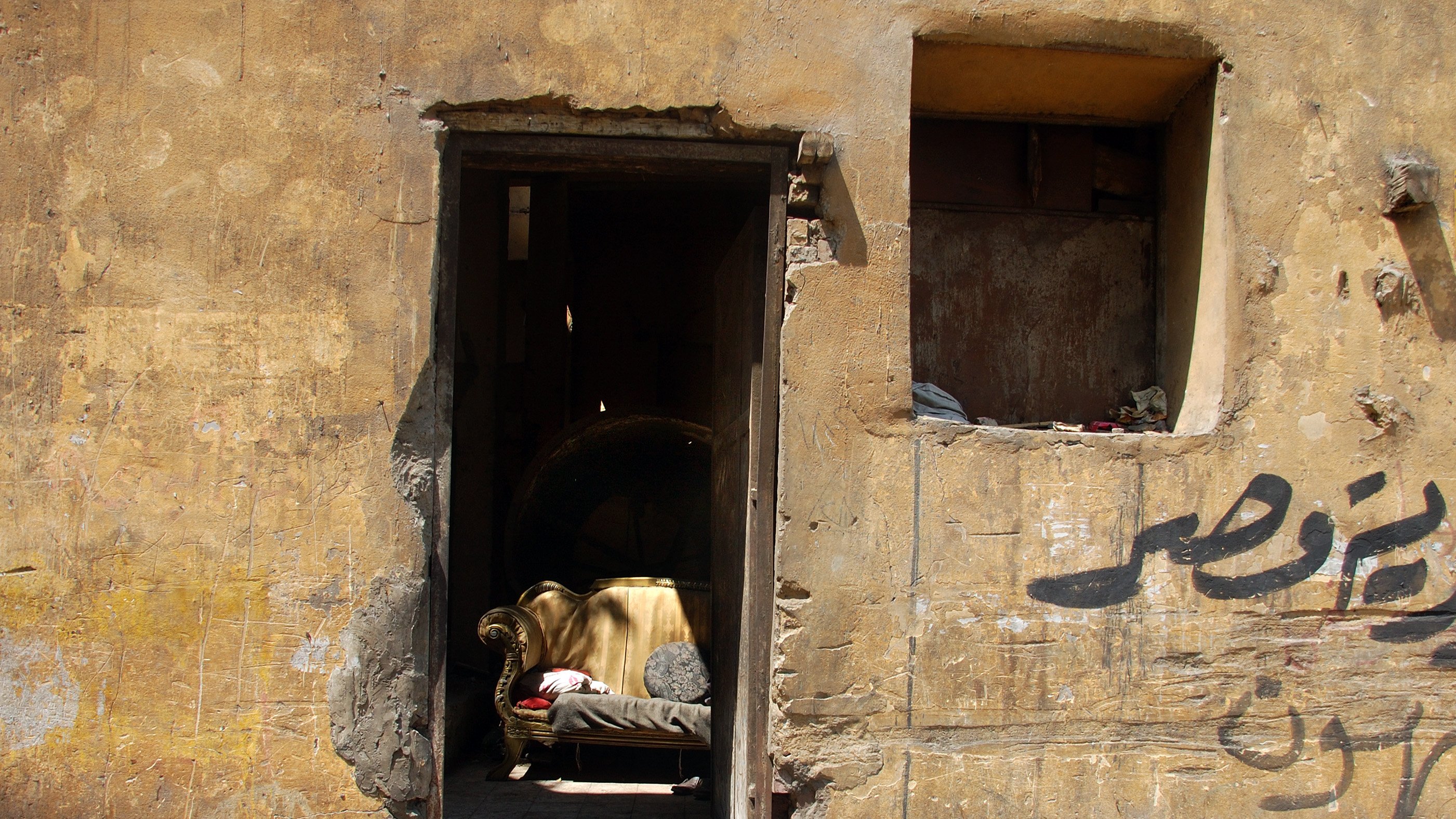 تصویر نمایی از یک درب ورودی به یک ساختمان قدیمی است که دیوارهای آن رنگ و بافت زرد و کهنه‌ای دارند. در وسط در، یک کاناپه قدیمی و ساده قرار دارد که به نظر می‌رسد سال‌ها بدون استفاده مانده است. در سمت چپ تصویر، بخشی از دیوار به صورت تخریب شده دیده می‌شود و یک پنجره کوچک بالای درب نیز وجود دارد که نیمی از آن به طور کامل تخریب شده است. روی دیوار نوشته‌هایی به زبان عربی دیده می‌شود که پس‌زمینه‌ای از فضای شهری و قدیم را به تصویر می‌کشد. کل صحنه حس نوستالژیک و متروکه‌ای را منتقل می‌کند.