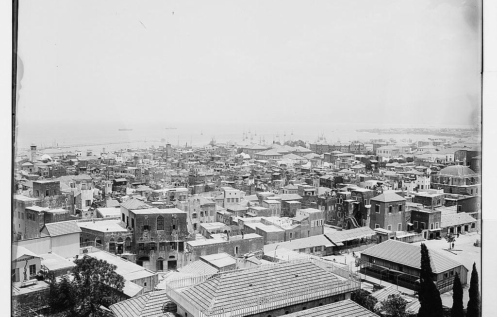 L'image présente une vue panoramique d'une ville côtière. On peut voir de nombreux bâtiments aux toits en tôle, ainsi que des structures en maçonnerie. La mer est visible à l'arrière-plan, avec quelques bateaux amarrés. Les rues semblent étroites et bien dessinées, et il y a des arbres éparpillés dans la zone. L'ensemble donne une impression de densité urbaine et de vie maritime. La photographie semble ancienne, capturant l'architecture et l'ambiance d'une époque révolue.