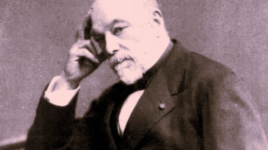 L'image montre un homme mature assis, avec une expression pensive. Il porte un costume sombre et une cravate. Sa main droite touche son menton, ce qui donne une impression de réflexion. Le fond est sombre, ce qui fait ressortir le personnage. La photographie a un style ancien, typique des portraits du 19ème siècle.