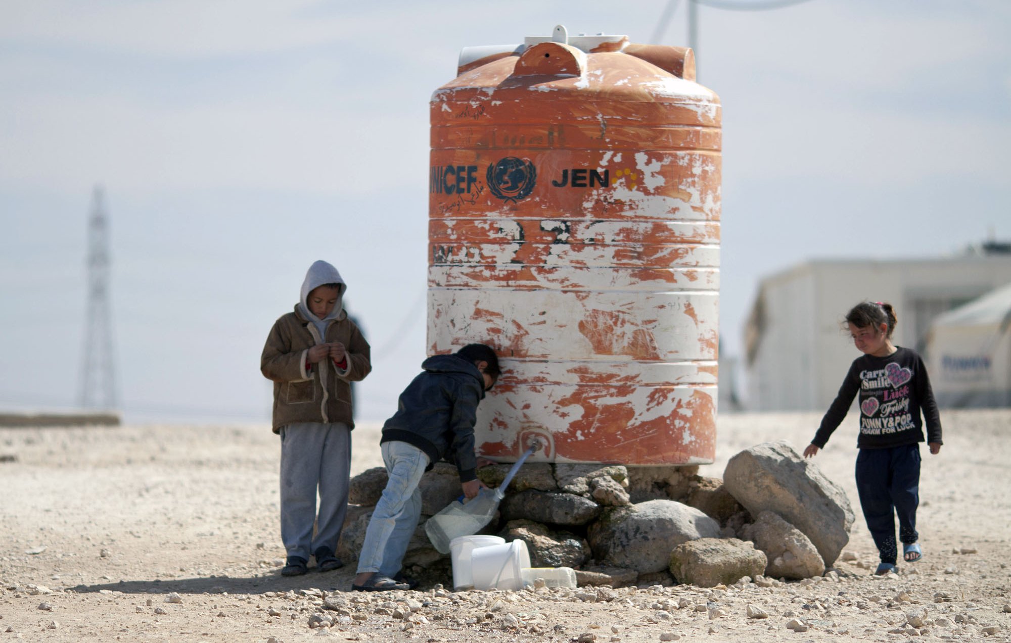 The image depicts children interacting near a large water tank. One child appears to be filling a container with water from a tap on the tank, while another child stands beside them. A third child is standing apart, looking at their phone or device. The water tank is orange and white, marked with a logo that indicates it is associated with a humanitarian organization. The surrounding area is dry and rocky, suggesting a challenging environment.
