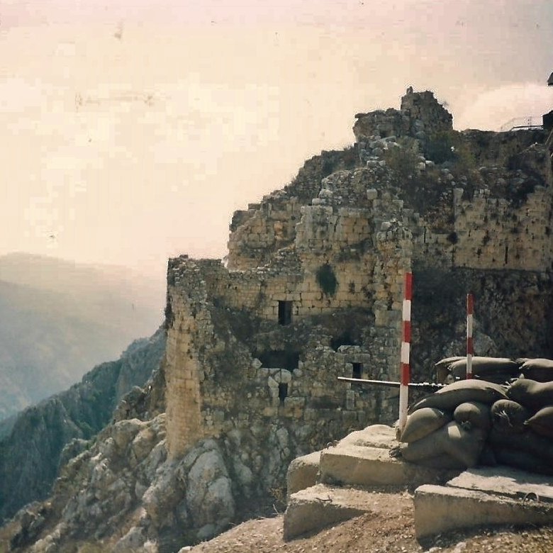 L'image montre les ruines d'une vieille forteresse perchée sur une colline, avec des murs en pierre partiellement érodés. Des drapeaux flottent au sommet de la structure. En premier plan, on peut voir des sacs de terre disposés en protection, suggérant une utilisation militaire. La vaste vallée et les montagnes se dessinent à l'arrière-plan, créant une ambiance de paysages montagneux majestueux. L'ensemble évoque une atmosphère historique et stratégique.