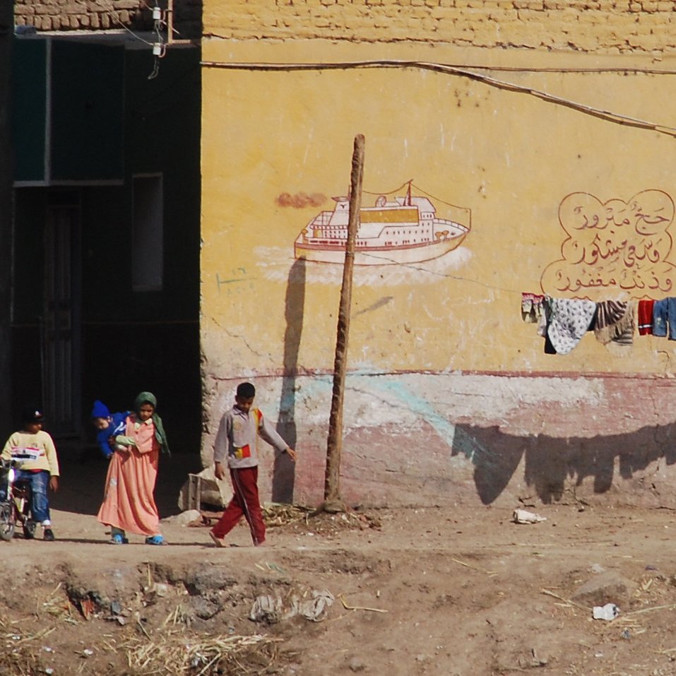 The image depicts a scene from a rural or semi-urban area, featuring a group of people interacting outside. A child in a wheelchair is being pushed forward by an adult, while two other children walk beside them. In the background, there is a building with a yellow wall that has some painted decorations or text. Above this, a clothesline displays various articles of clothing hanging to dry. The ground appears uneven, with some debris scattered around. Overall, the scene conveys everyday life in a community setting.
