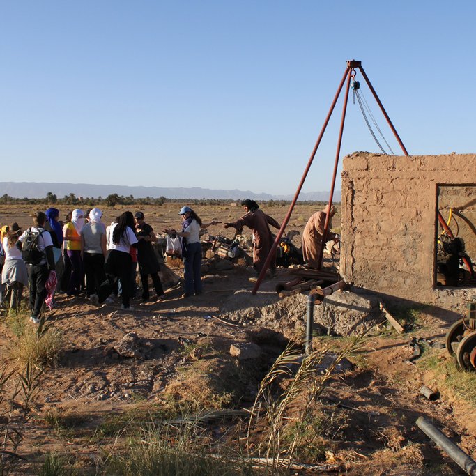 L'image montre un groupe de personnes rassemblées autour d'une structure en terre, possiblement une ancienne construction ou un puits. Il y a une machine ou un mécanisme visible à l'intérieur de la structure. L'environnement est désertique, avec un sol aride et des montagnes en arrière-plan. Le ciel est dégagé, reflétant une ambiance ensoleillée. Les participants semblent s'intéresser à l'activité en cours, observant attentivement.