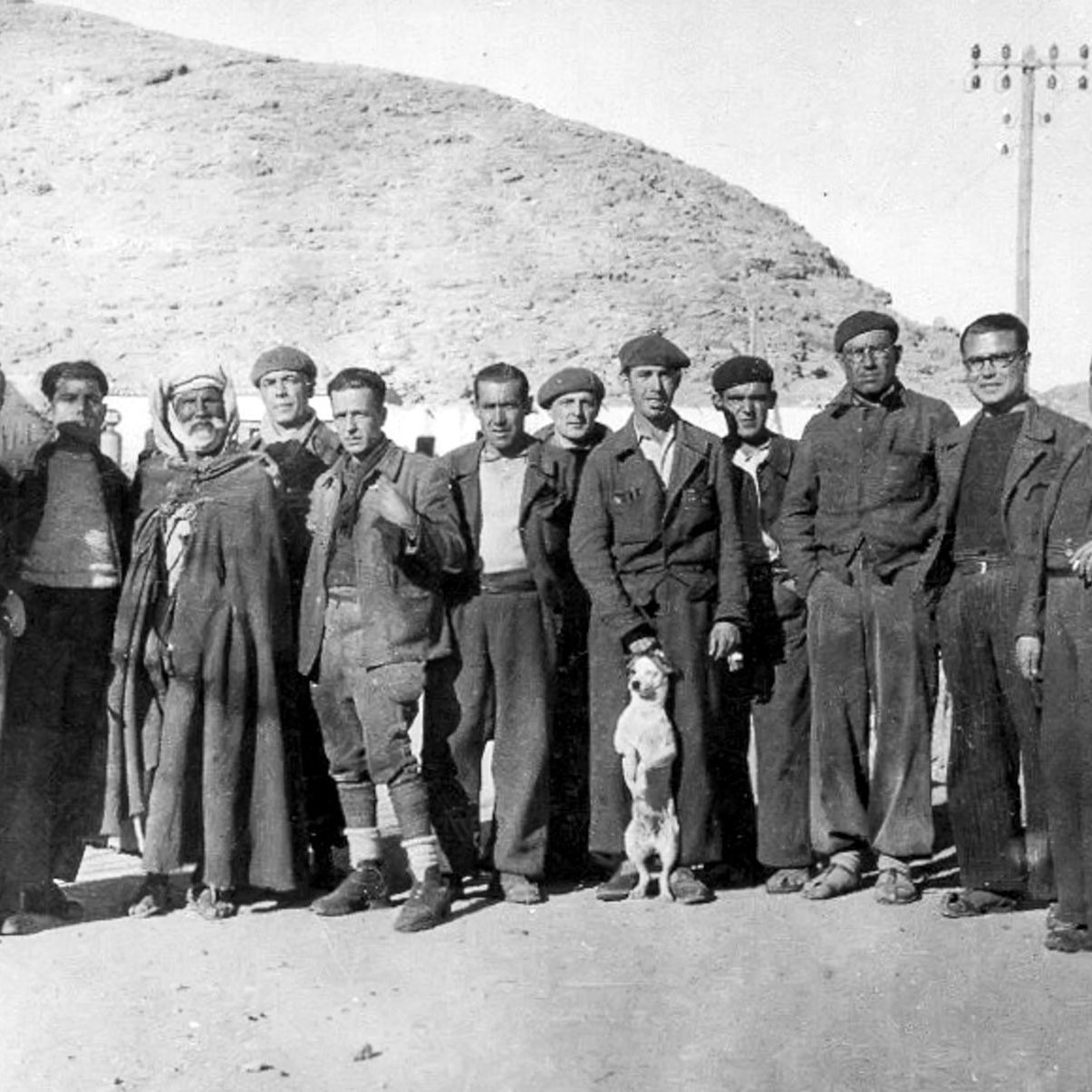 L'image en noir et blanc montre un groupe d'hommes se tenant debout ensemble, probablement dans un environnement rural ou montagneux. Ils portent des vêtements qui semblent être des tenues de travail ou militaires. Au centre, un homme en costume traditionnel se distingue des autres. Un chien se trouve également parmi le groupe. Les hommes affichent diverses expressions, allant des sourires aux regards sérieux. L'arrière-plan présente des collines, et quelques bâtiments peuvent être aperçus. L'ensemble évoque une période historique, probablement au début ou au milieu du 20e siècle.