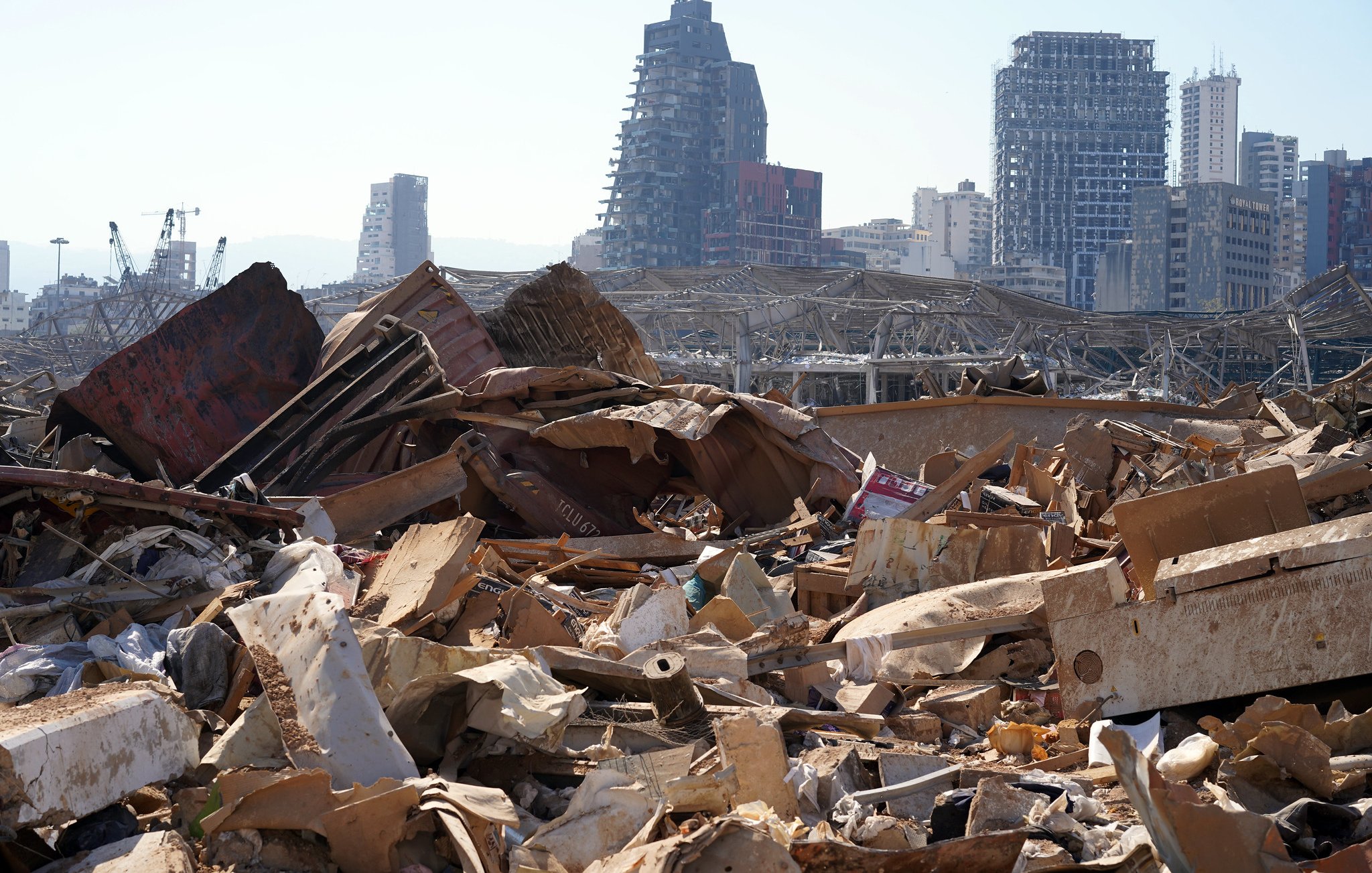 L'image montre un paysage urbain dévasté, avec des débris et des matériaux en désordre dans le premier plan. On peut voir des bâtiments partiellement détruits en arrière-plan, témoignant des dommages importants. L'atmosphère semble désolée, illustrant l'impact d'une catastrophe, probablement une explosion ou une destruction massive. Des structures modernes sont visibles, mais leur état montre les effets néfastes de l'événement. Le ciel est dégagé, contrastant avec la gravité de la scène au sol.