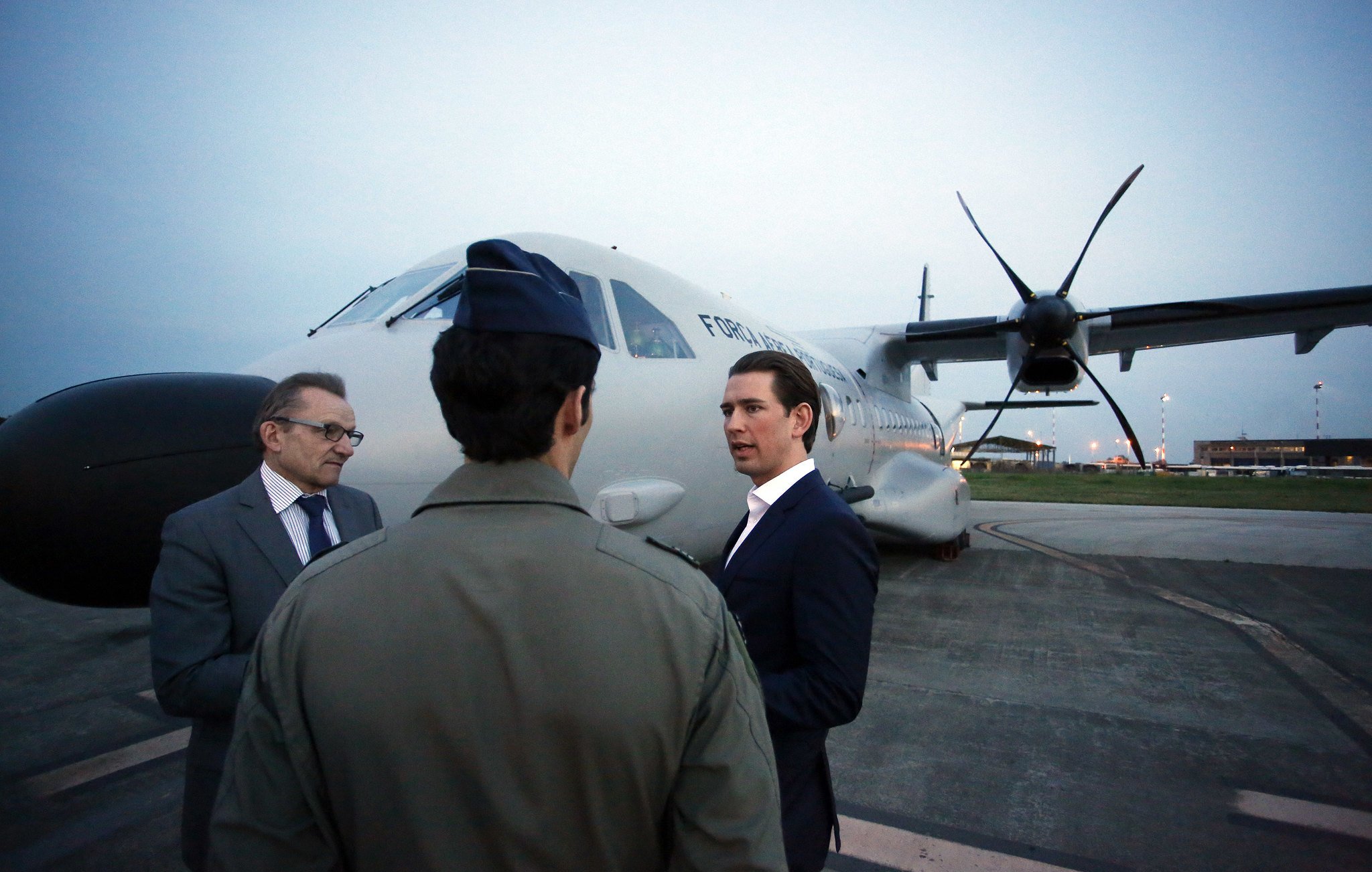 L'image montre trois hommes près d'un avion. L'un des hommes porte un uniforme militaire, tandis que les deux autres sont habillés dans des costumes. Ils semblent être en conversation, face à l'avion de transport, qui est stationné sur le tarmac à la tombée de la nuit. L'éclairage ambiant est doux, et on peut voir des lumières à l'arrière-plan, probablement d'un aéroport ou d'une base aérienne.