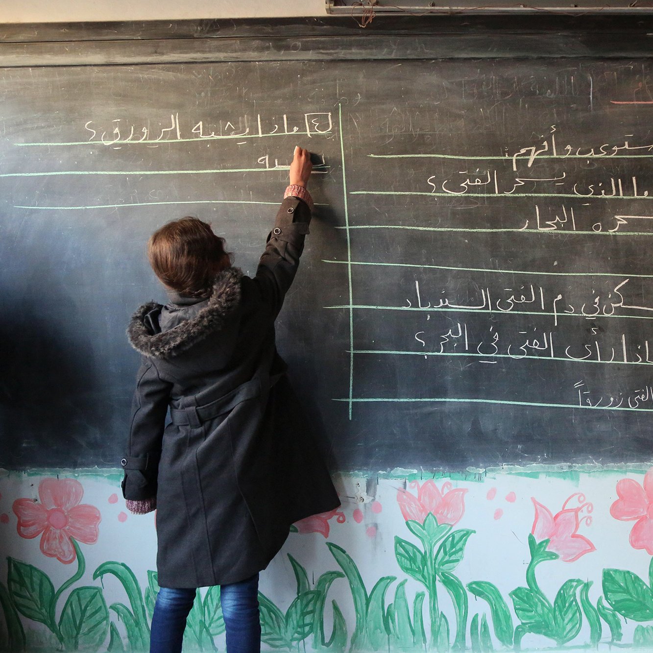 The image depicts a classroom scene where a student is writing on a blackboard. The blackboard has lines of text in Arabic, while the bottom of the board is decorated with colorful flower drawings. The student, wearing a coat, is focused on writing, and there are other individuals in the background, potentially teachers or classmates. The setting appears to be an educational environment.