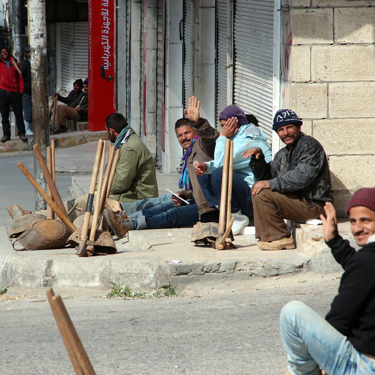 The image depicts a group of men sitting on a sidewalk in an urban setting. They appear to be relaxed, some waving at the camera, and are dressed in casual clothing. The street is relatively quiet, with a few vehicles in the background. There are also some construction tools and materials scattered around, suggesting they may be waiting for work or gathering. The environment looks somewhat desolate, with shuttered storefronts and a hint of a dusty road.