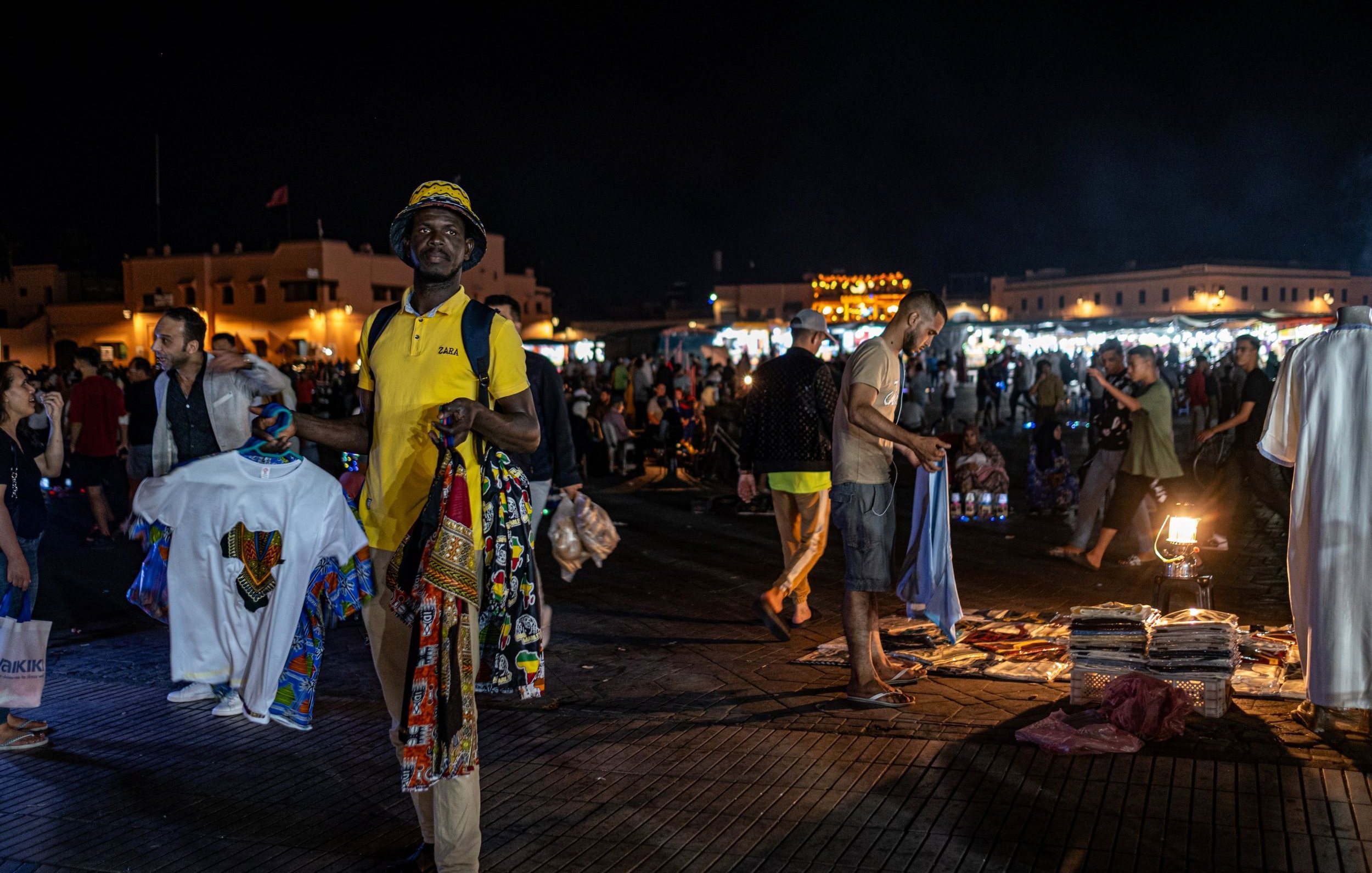 L'image montre un marché nocturne animé, probablement dans un endroit comme Marrakech. On peut voir des gens se déplacer dans une ambiance vibrante. Au premier plan, un homme porte un t-shirt jaune et tient plusieurs vêtements traditionnels colorés. En arrière-plan, d'autres personnes flânent et explorent les étals, tandis que des lumières scintillantes créent une atmosphère festive. Des tapis et d'autres articles sont visibles au sol, ajoutant à l'attrait du marché.