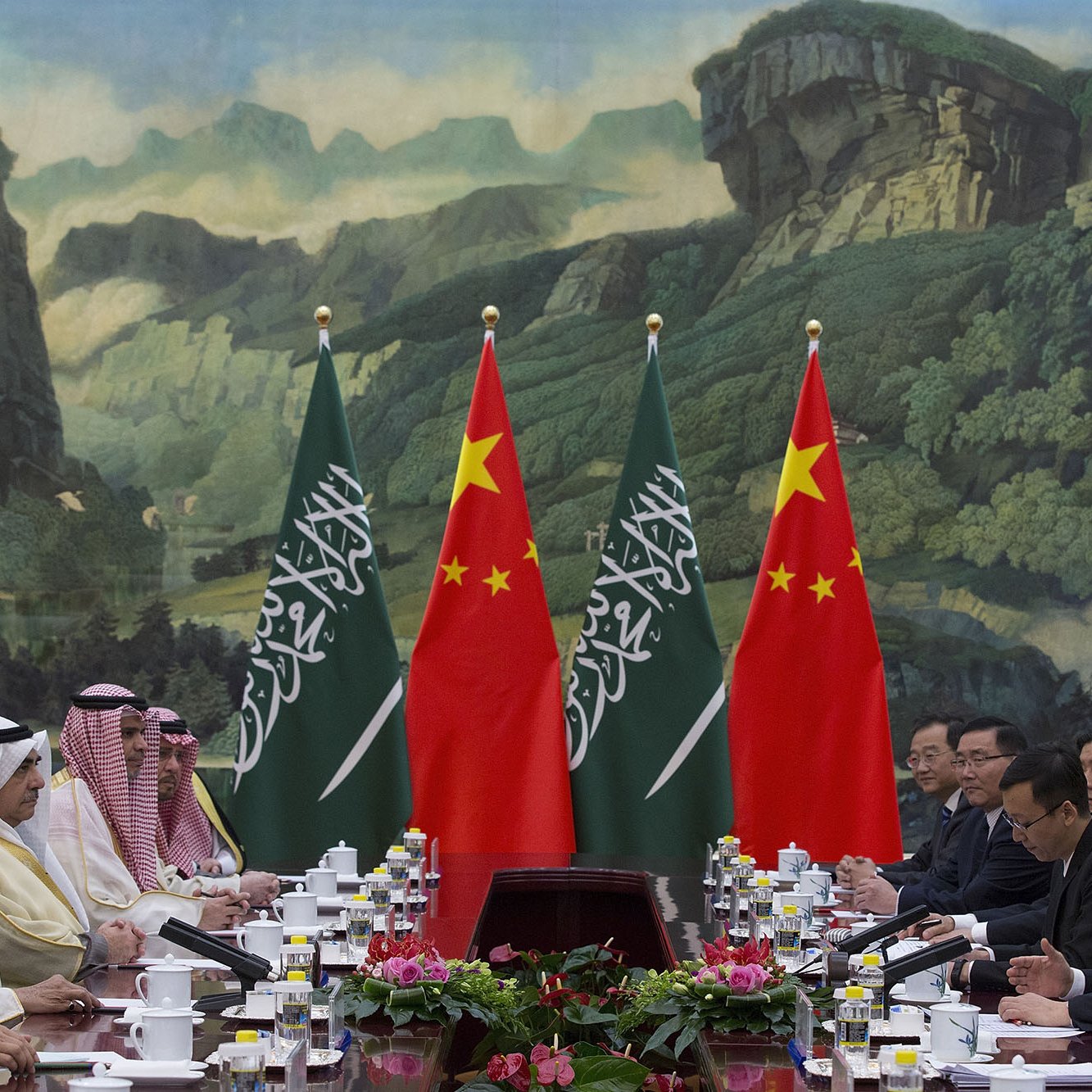 L'image montre une réunion diplomatique entre des représentants de l'Arabie saoudite et de la Chine. Les participants sont assis à une table, entourés de drapeaux des deux pays. En arrière-plan, on peut voir une grande fresque représentant des paysages naturels. La scène dégage une ambiance formelle, typique des sommets ou des négociations internationales. Sur la table, il y a des documents et des tasses de thé.