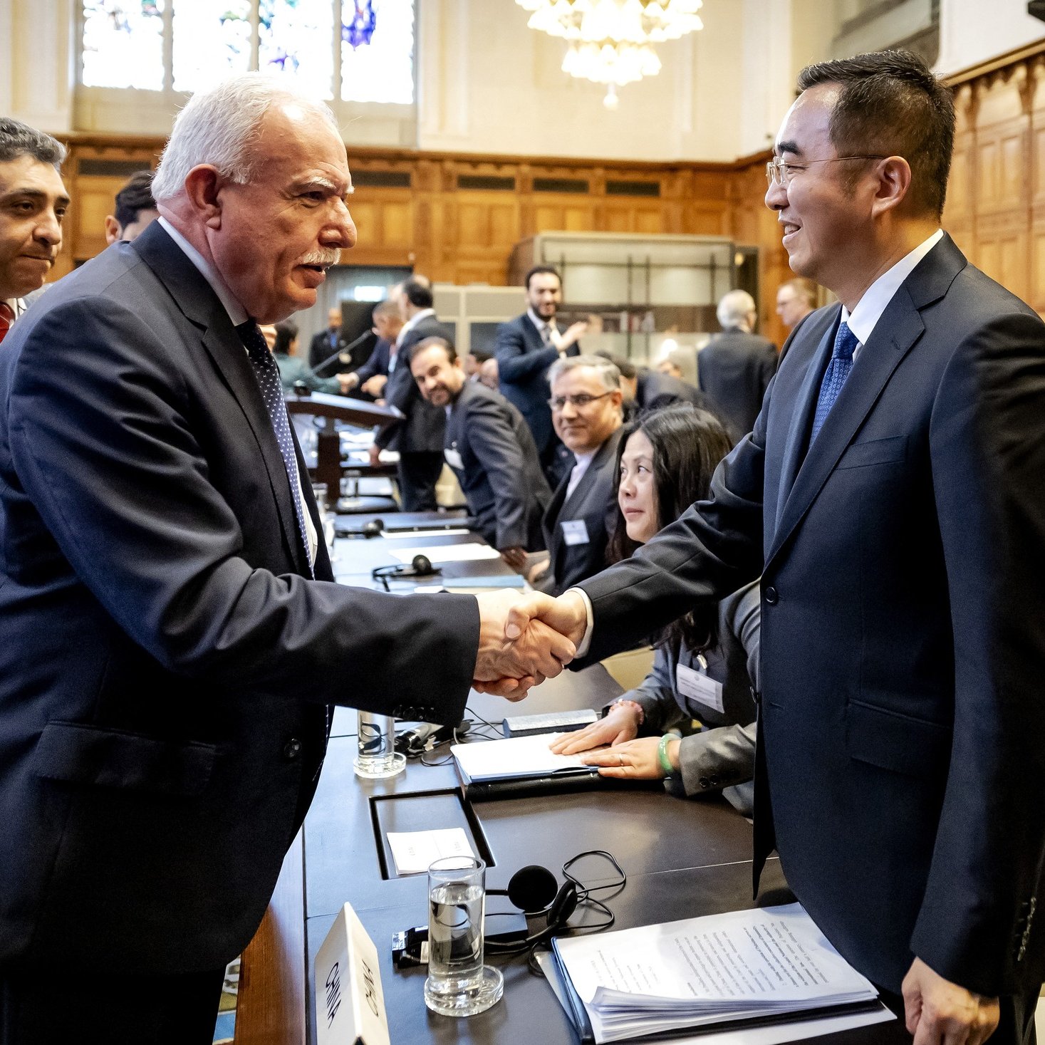 الصورة تظهر مشهدًا من اجتماع رسمي، حيث يتم تبادل المصافحات بين شخصين يجلسان على طاولة. الشخص على اليسار يبدو أنه يتحدث ويبتسم، بينما الشخص على اليمين يقوم بمصافحته بيديه. في الخلفية، يوجد عدد من الأشخاص الآخرين الذين يجلسون على الطاولة أو يقفون، ويبدو عليهم الاستماع أو المشاركة في الحوار. المكان يبدو أنيقًا، مع تصميم داخلي جميل يشمل نوافذ كبيرة وإضاءة جيدة.