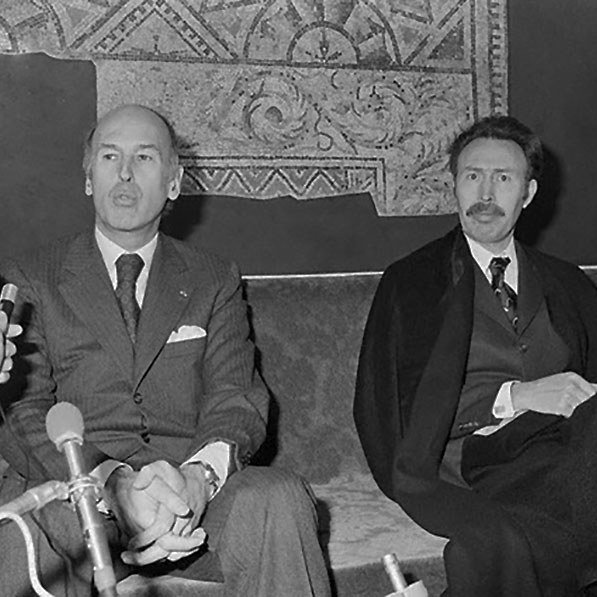 L'image montre deux hommes assis sur un canapé, apparemment lors d'une conférence de presse ou d'une rencontre. Ils semblent sérieux et attentifs, avec des microphones en face d'eux, indiquant qu'ils sont en train de répondre à des questions. L'un des hommes porte un costume sombre et a une expression réfléchie, tandis que l'autre, à sa droite, est vêtu d'une façon plus formelle, peut-être avec une écharpe ou un manteau. En arrière-plan, il y a un motif mural qui ajoute une certaine ambiance à la scène.