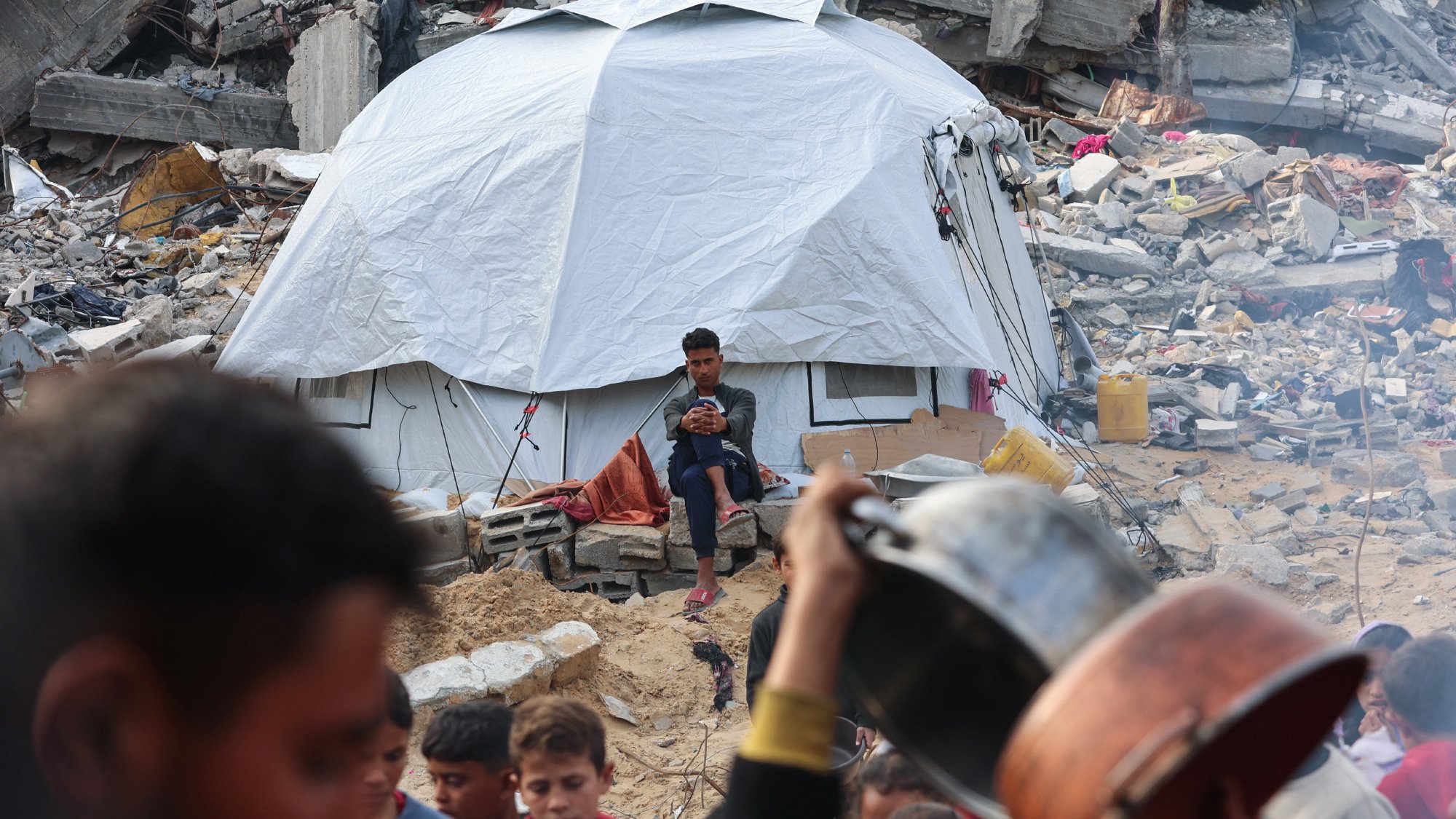 در این تصویر، یک چادر سفید در میان خرابهها قرار دارد و یک نوجوان در حال نشستن و استفاده از گوشی تلفن همراه است. در جلوی چادر، یک گروه از کودکان و افراد بزرگسال دیده میشوند که به سمت دوربین نگاه میکنند یا در حال انجام کارهایی هستند. زبالهها و خرابههای ساختمانهای تخریبشده در پسزمینه به وضوح احساس ناامیدی و بحران را منتقل میکنند.