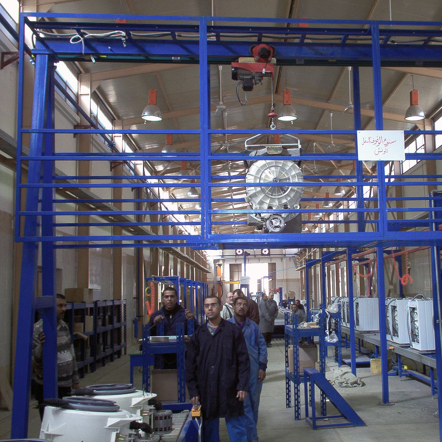 الصورة تبين مشهداً داخل مصنع أو ورشة عمل. يظهر في الصورة عدة أشخاص يعملون في بيئة صناعية. توجد آلات وأجهزة كهربائية مرتبة على طاولات أو أسطح عمل، بالإضافة إلى نظام تعليقي يحمل بعض المعدات في السقف. الإضاءة جيدة، مما يساهم في خلق جو عمل مناسب. كما يوجد لافتة معلقة على الجدار تحمل نصاً، مما يشير إلى وجود توجيهات أو معلومات مهمة في المكان.