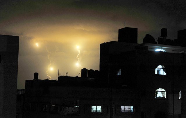 L'image montre un ciel nocturne illuminé par des éclairs, avec des nuages sombres en arrière-plan. On peut voir des bâtiments en silhouette, avec des fenêtres lumineuses qui créent un contraste avec l'obscurité environnante. Les éclairs apparaissent comme des points lumineux dans le ciel, ajoutant une atmosphère dramatique à la scène.