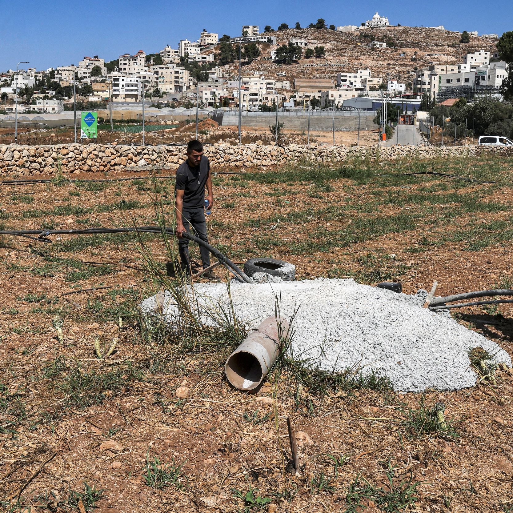 L'image montre deux hommes travaillant sur un terrain en plein air, probablement dans une zone rurale. Ils manipulent des tuyaux en plastique et se trouvent à proximité d'un tas de ciment ou de béton, ainsi qu'un petit tuyau qui sort du sol. À l'arrière-plan, on peut apercevoir des bâtiments et des collines. La végétation est clairsemée, et le sol semble fraîchement labouré ou non entretenu.