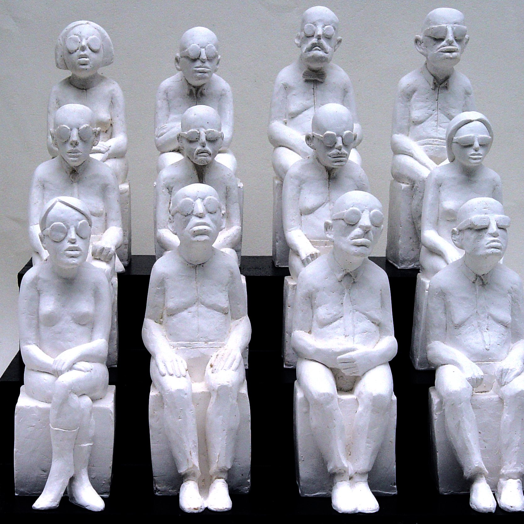 L'image représente une sculpture où plusieurs figures humaines, toutes de couleur blanche, sont assises sur des bancs. Les personnages sont stylisés avec des traits simples et géométriques, portant des lunettes, et semblent avoir des expressions neutres ou pensive. Ils sont agencés en un groupe de dix, disposés en plusieurs rangées, créant une impression d'unité et de contemplation. Les sculptures semblent évoquer des thèmes tels que la solitude, la réflexion ou la condition humaine.