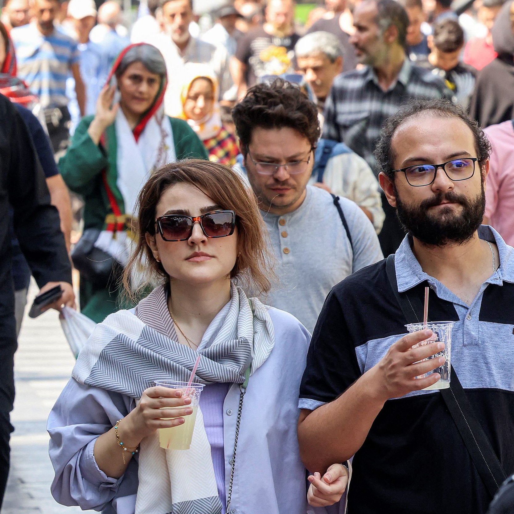 L'image montre une scène animée d'une rue où plusieurs personnes marchent. Au premier plan, deux personnes se déplacent côte à côte. Un homme portant des lunettes et une chemise à rayures tient un verre. À côté de lui, une femme avec des lunettes de soleil et une écharpe semble s'engager dans une conversation. En arrière-plan, d'autres personnes marchent, créant une atmosphère dynamique. On peut voir une variété de styles vestimentaires, suggérant une diversité culturelle.