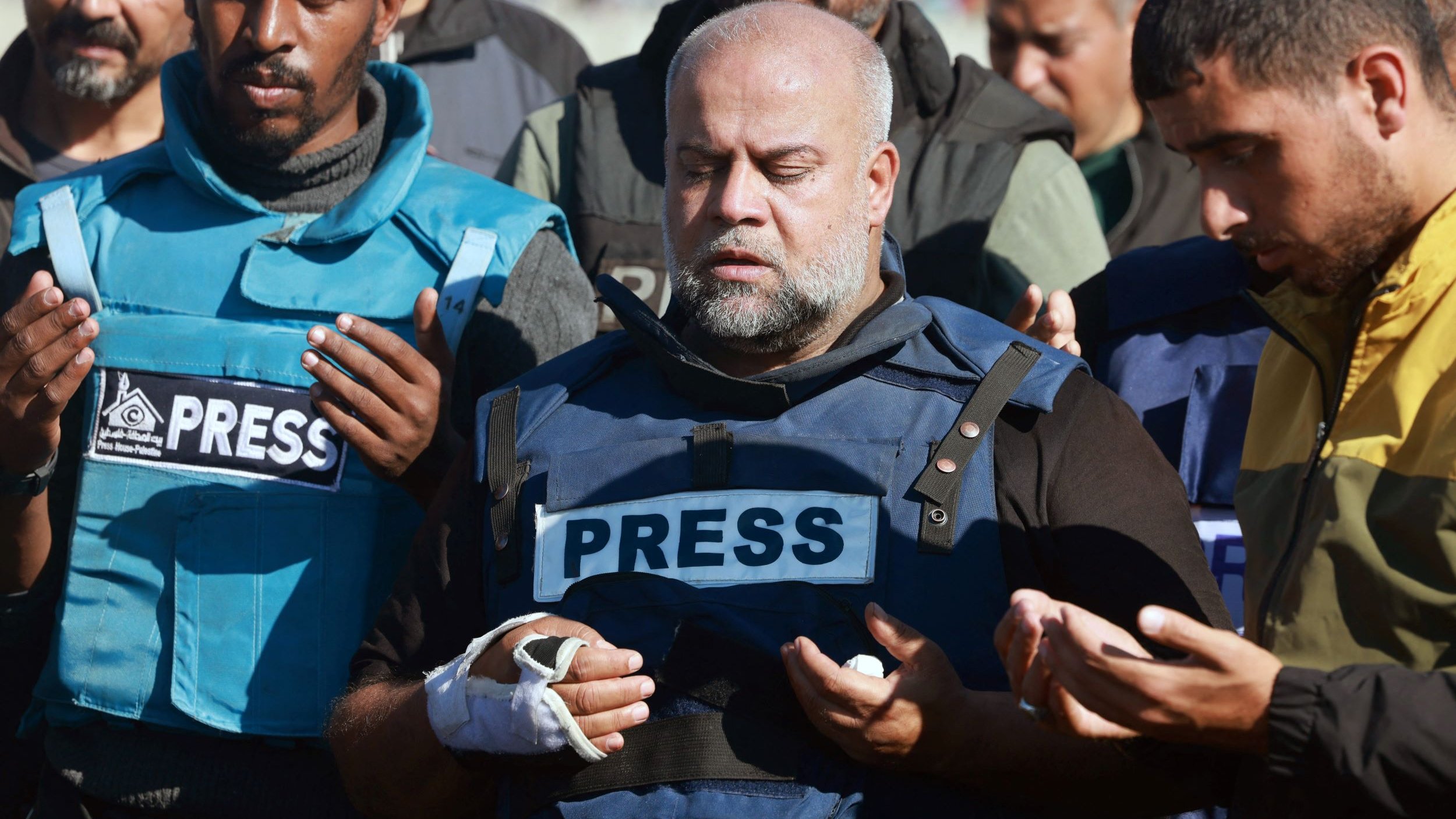 L'immagine mostra un gruppo di persone in piedi che sembrano partecipare a un momento di preghiera o riflessione. In primo piano, un uomo indossa un giubbotto con la scritta "PRESS" ed ha una fasciatura su un braccio. Gli altri membri del gruppo, che indossano abbigliamento simile, appare coinvolti nell'atto di pregare, con le mani sollevate. L'atmosfera sembra essere solenne e carica di emotività.