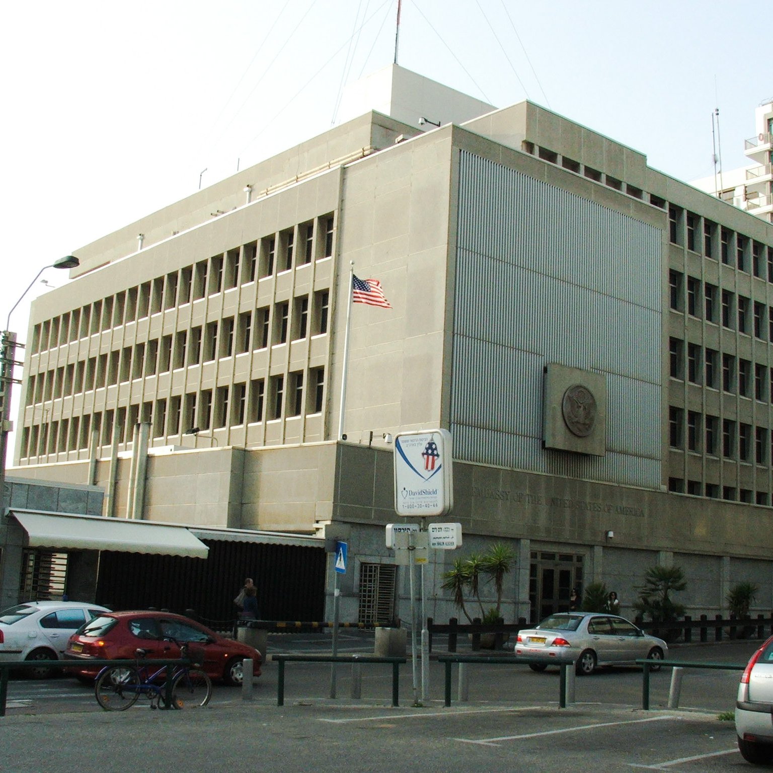 L'image montre un bâtiment moderne avec une architecture fonctionnelle. Il est en béton, avec plusieurs fenêtres rectangulaires sur la façade. Un drapeau américain est visible sur le toit, indiquant que c'est une représentation diplomatique. À l'avant du bâtiment, il y a des panneaux de signalisation et quelques véhicules garés le long de la route. L'environnement semble urbain, avec d'autres immeubles en arrière-plan.