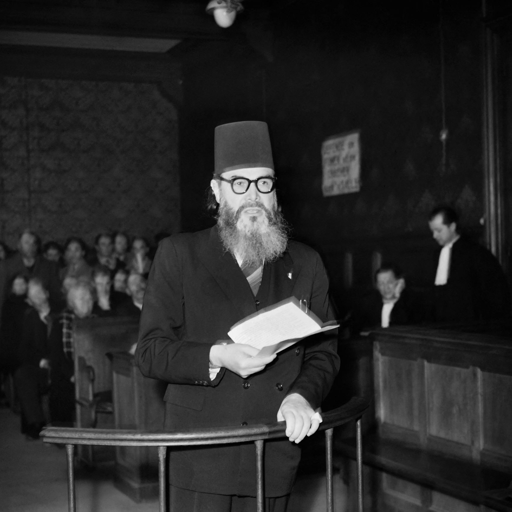 L'image montre un homme portant un turban noir et des lunettes, se tenant debout dans une salle d'audience. Il lit un document, tandis qu'un groupe de personnes l'écoute attentivement en arrière-plan. L'environnement est sombre, avec des boiseries et des bancs de juge visibles. Une personne en toge est également présente, ajoutant à l'atmosphère judiciaire de la scène.