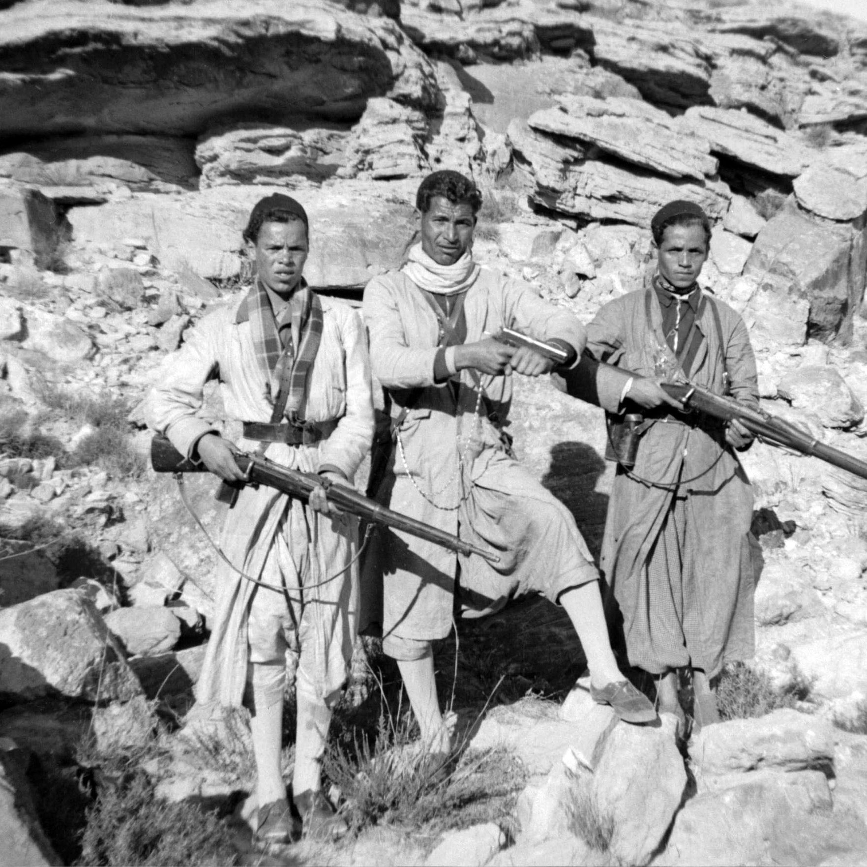 L'image montre trois hommes vêtus de vêtements traditionnels, tenant chacun un fusil. Ils se tiennent debout sur un sol rocheux, entourés de grandes pierres et de formations rocheuses. L'environnement semble austère et désertique. Les hommes ont des expressions sérieuses et sont alignés côte à côte. Leur posture et l'équipement qu'ils portent suggèrent qu'ils peuvent être des membres d'un groupe armé ou des guerriers appartenant à une communauté spécifique.
