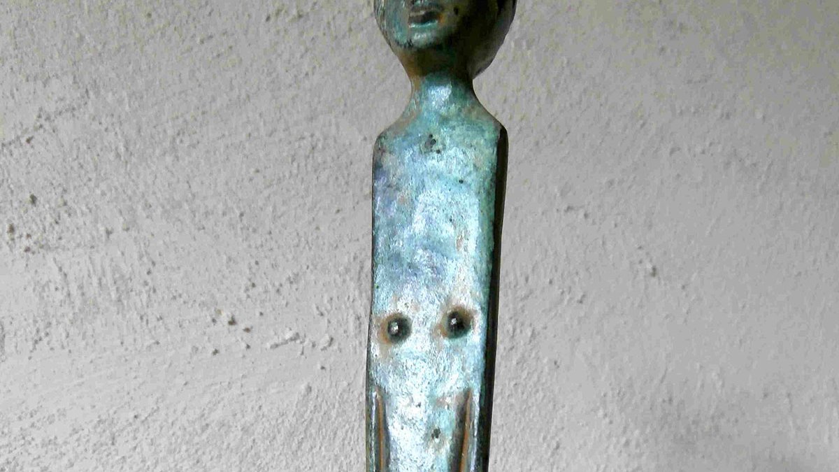 L'image montre un objet en métal, probablement en bronze, qui semble être une sorte de sculpture ou de barre. La partie supérieure représente une tête humaine stylisée, tandis que la tige qui descend présente des éléments qui pourraient évoquer un corps. La surface a une patine verdâtre, indiquant son ancienneté. L'ensemble de l'objet évoque une forme artistique, fusionnant des éléments figuratifs et abstraits.