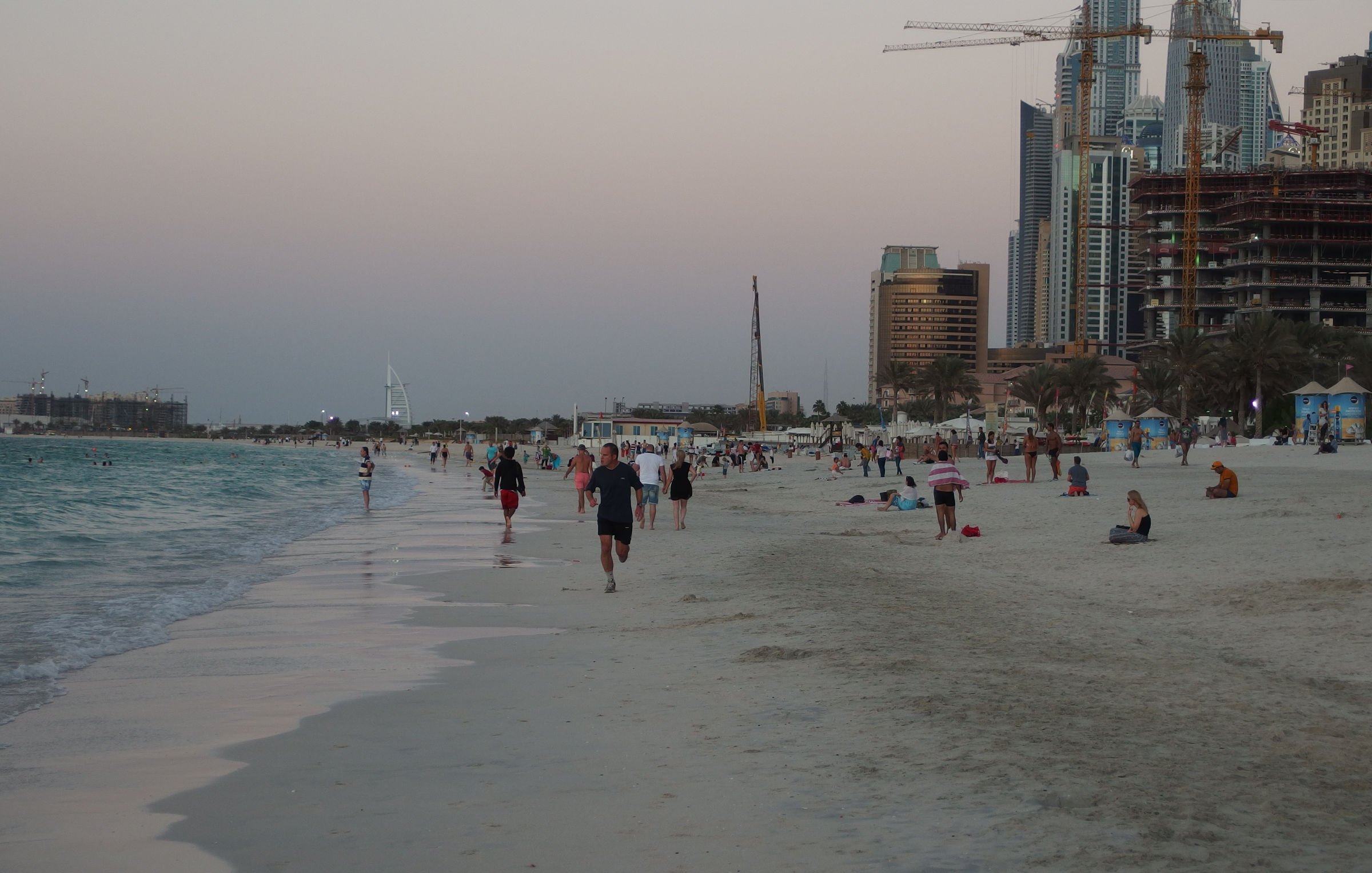 L'image montre une plage avec des personnes marchant le long du rivage. On aperçoit des baigneurs et des groupes assis sur le sable. En arrière-plan, on distingue des bâtiments modernes et des gratte-ciel, symboles d'une grande ville, ainsi que des grues témoignant de travaux de construction. Le ciel présente des teintes douces, probablement au moment du crépuscule, créant une atmosphère paisible et attractive. Les vagues de la mer semblent calmes, ajoutant à la sérénité du paysage.