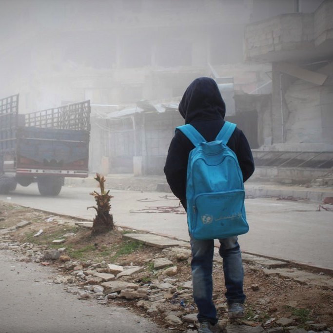 L'image montre un jeune garçon de dos, portant un grand sac à dos bleu. Il se tient sur une rue où la visibilité est réduite en raison d'un épais brouillard. En arrière-plan, on peut apercevoir des bâtiments partiellement détruits et un camion stationné. Le sol est jonché de débris et de petites plantes. L'atmosphère est sombre et mélancolique, évoquant une scène de désolation.