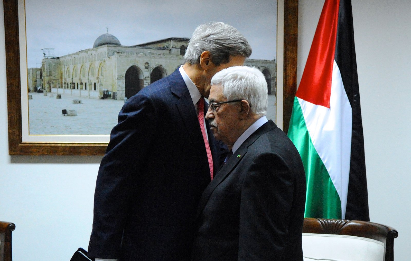L'image montre deux hommes en train de s'incliner l'un vers l'autre dans un environnement formel. En arrière-plan, il y a une grande photo d'un monument architectural, probablement une mosquée, et un drapeau représentant les couleurs rouge, noir, blanc et vert. Les hommes semblent avoir une conversation sérieuse, et l'un d'eux est habillé en costume, tandis que l'autre porte une tenue plus traditionnelle. L'atmosphère de la scène évoque un moment de négociation ou de discussion diplomatique.