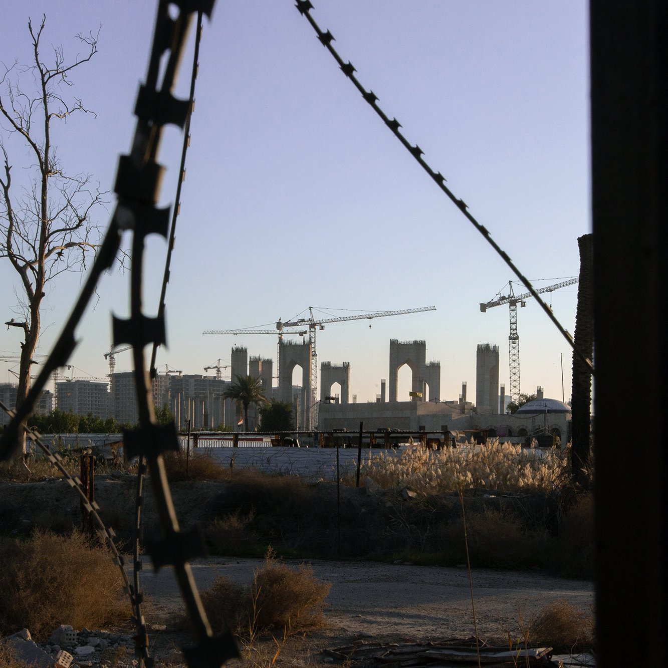 L'image présente un paysage urbain en transformation, avec des structures inachevées à l'arrière-plan, entourées de grues de construction. En premier plan, on peut voir des barbelés et des éléments de végétation desséchée, suggérant un environnement abandonné ou en déclin. La lumière douce du crépuscule ajoute une ambiance mélancolique à la scène, mettant en contraste l'espoir de la construction avec la désolation du terrain environnant.