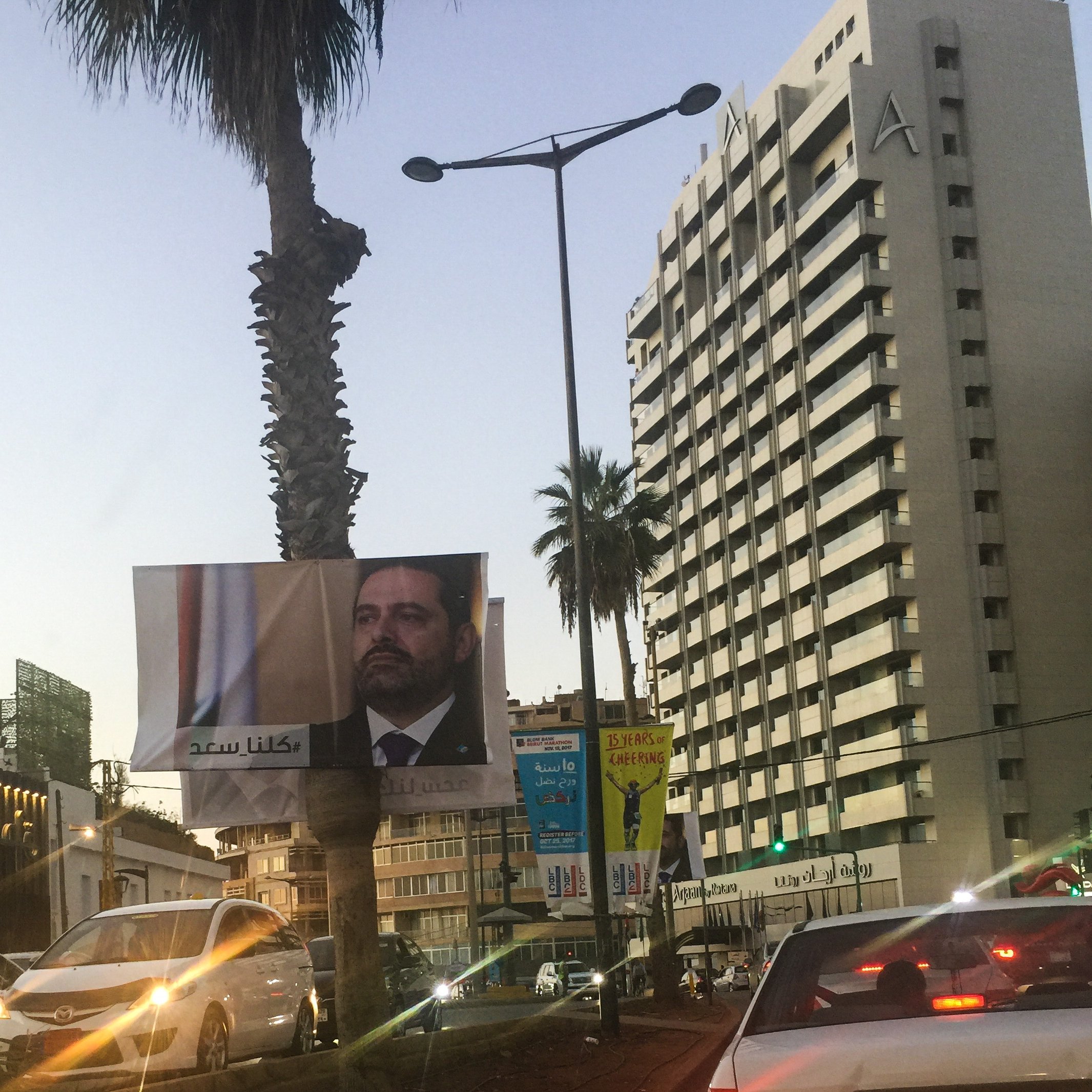 در این تصویر یک بیلبورد بزرگ در کنار خیابان دیده می‌شود که چهره یک مرد را نمایش می‌دهد. او نسبت به دوربین نگاهی جدی دارد و به نظر می‌رسد که یک شخصیت سیاسی باشد. در زمینه، ساختمان‌های بلند و درختان نخل دیده می‌شوند که فضای شهری را تشکیل می‌دهند. نور غروب خورشید بر روی صحنه افکنده شده که به آن حال و هوای خاصی می‌بخشد. چندین خودرو نیز در خط خیابان در حال حرکت هستند.