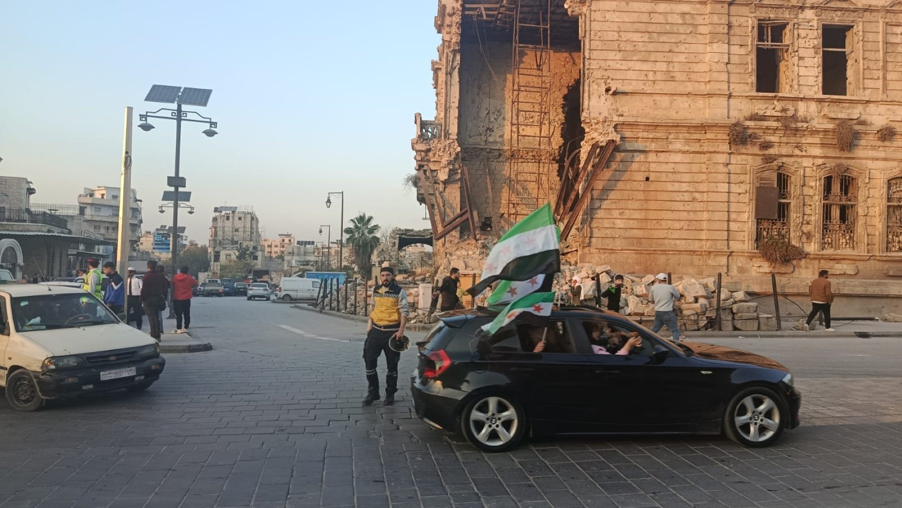 Una strada con una costruzione danneggiata, persone e un'auto con una bandiera.