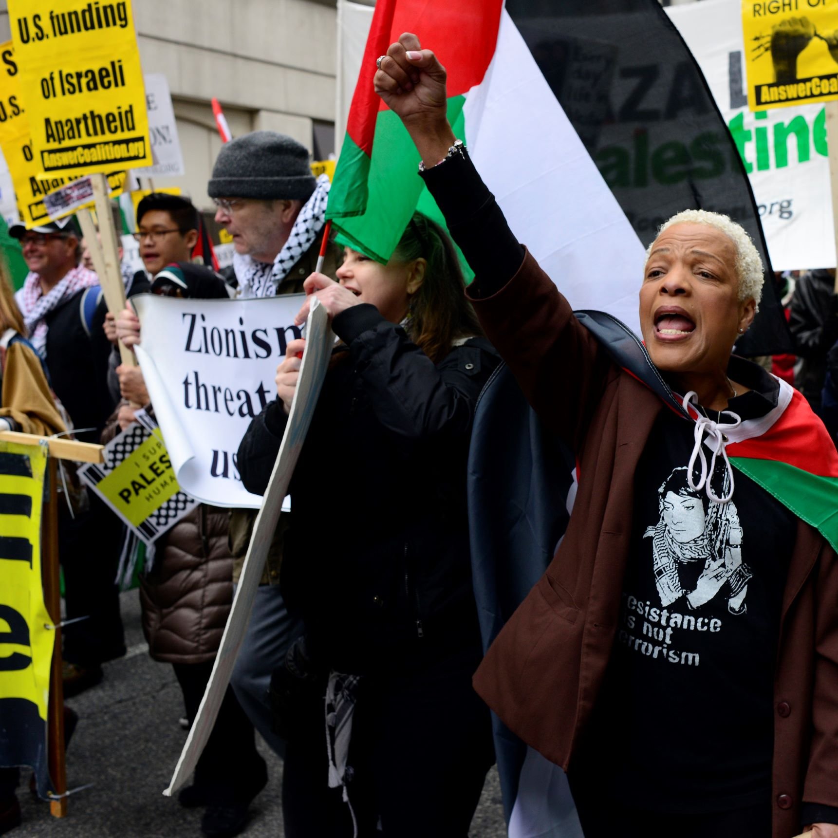 در این تصویر، گروهی از معترضان در یک راهپیمایی را مشاهده می‌کنیم. یک زن با پوست تیره در پیش‌زمینه قرار دارد که با فریاد و دست بالای خود، در حال بیان اعتراضاتش است. او پرچم‌های فلسطینی و نمادهای حمایت از حقوق فلسطینی‌ها را در دست دارد. دیگر افراد نیز در پس‌زمینه با پلاکاردهایی در حمایت از فلسطین و علیه صهیونیسم ایستاده‌اند. فضای عمومی احساس قوی از همبستگی و اعتراض را منتقل می‌کند.