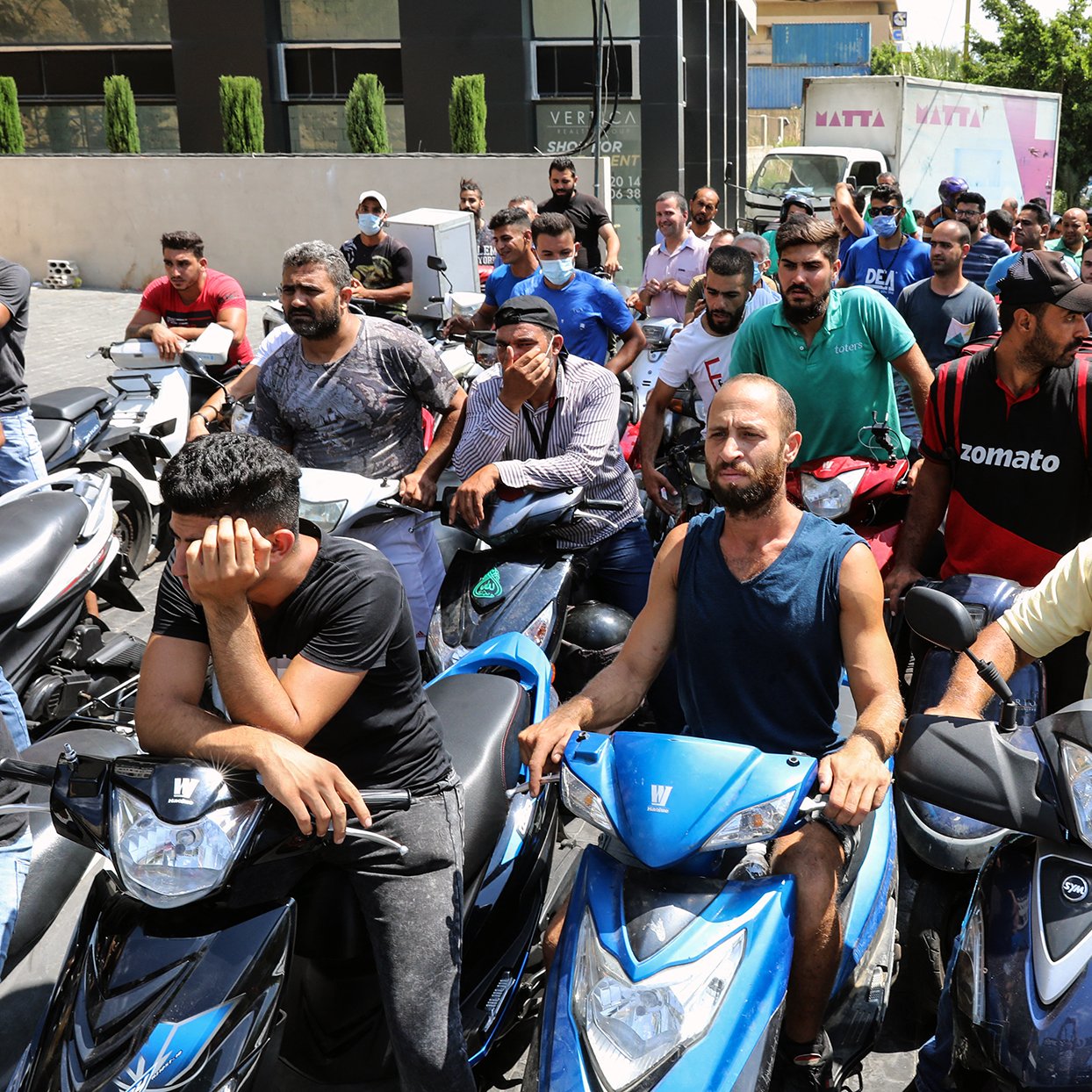L'image montre un groupe de personnes, principalement des hommes, rassemblés sur des motos dans une file d'attente. Ils semblent attendre pour quelque chose, peut-être un service ou un approvisionnement. Certains affichent des expressions de frustration ou d'impatience. En arrière-plan, on peut voir des bâtiments et des arbres qui ajoutent au cadre urbain. L'atmosphère donne une impression de tension liée à une situation d'attente prolongée.