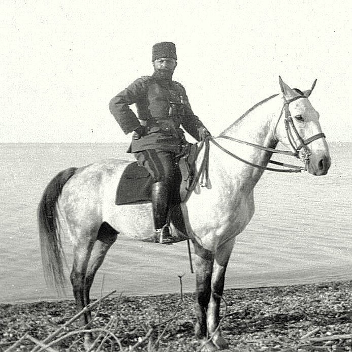 L'image montre un homme en uniforme militaire monté sur un cheval blanc. Il se tient debout sur la plage, près d'une étendue d'eau. L'homme porte un couvre-chef distinctif, et son uniforme semble traditionnel. Le paysage est simple, avec des galets au premier plan et un horizon calme en arrière-plan. L'atmosphère donne une impression de sérénité et de majesté.